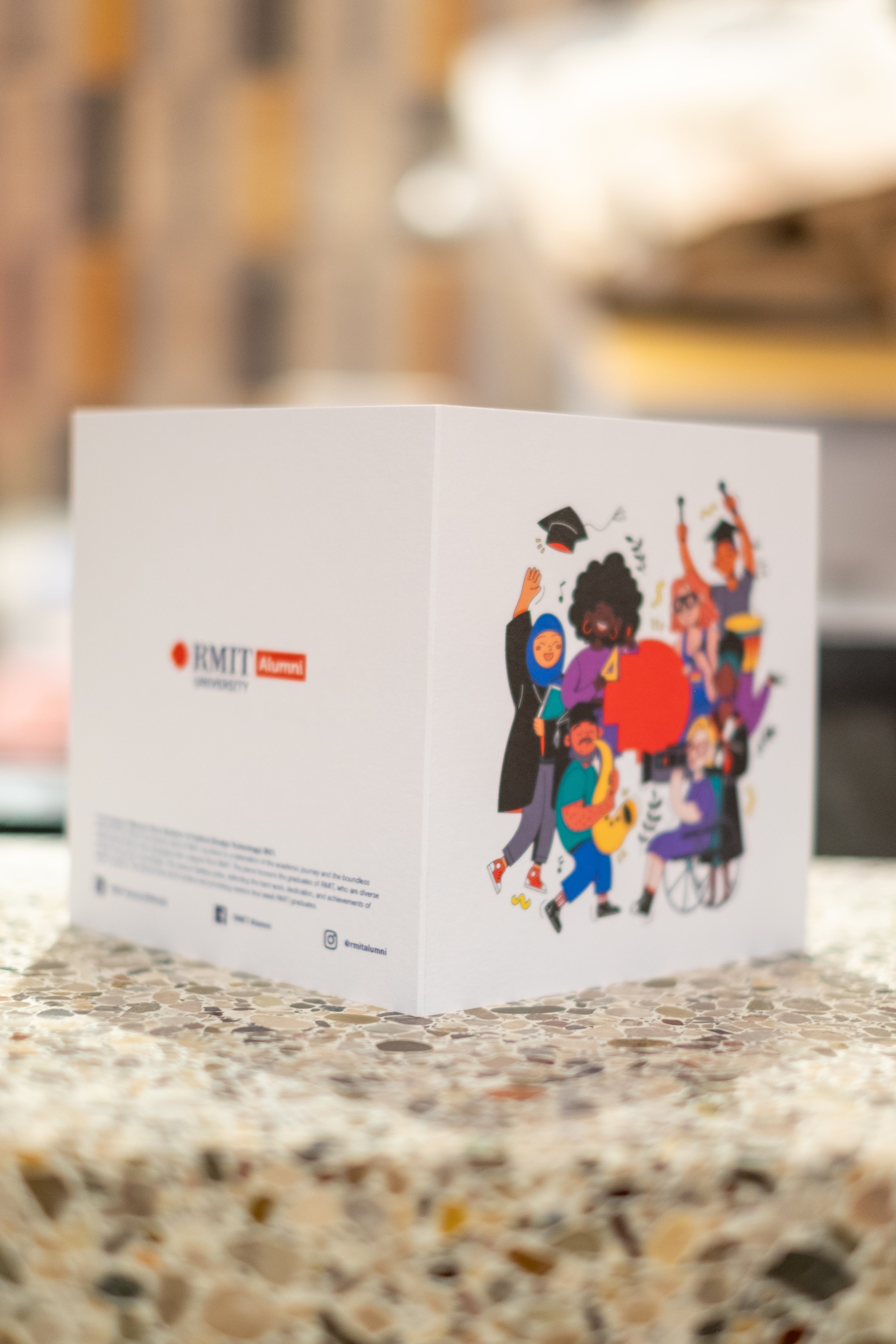 Slightly open full view of the alumni graduation card sitting on a light tabletop. The right side features a pattern of alumni-related icons, while the left side displays the RMIT University Alumni logo in navy and red, available at the RMIT Store.