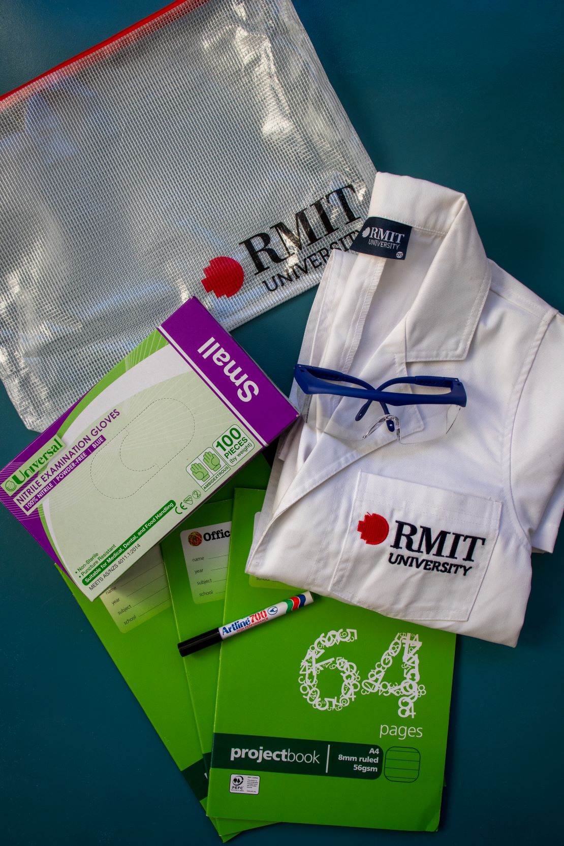 Contents of the RMIT Store Applied Science Kit including an embroidered RMIT branded lab coat, nitrile examination gloves, safety glasses, Artline 700 marker, project notebooks, and an RMIT-branded pouch, all arranged on a blue surface.
