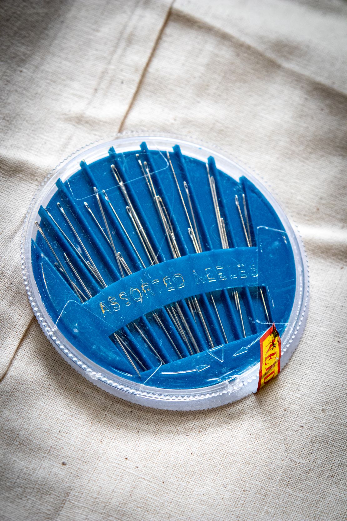 RMIT Store set of 30 assorted hand sewing needles in a round blue plastic case against a beige fabric backdrop, ideal for various sewing projects. The needles are neatly organised for easy selection and accessibility.