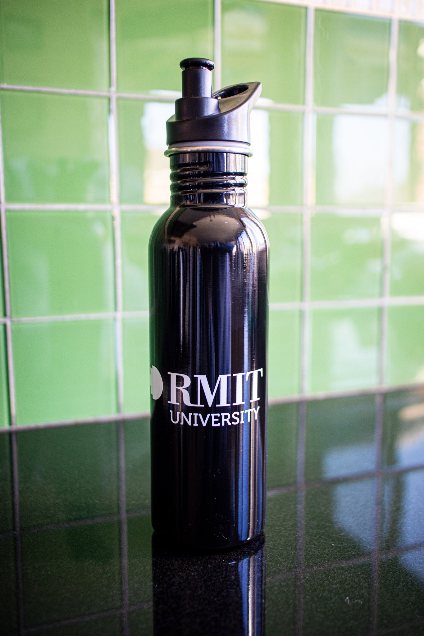 Full view of the black RMIT bottle with a white RMIT University logo centered on the body, black cap securely on the bottle, placed on a black tabletop with a green background. Available at the RMIT Store.