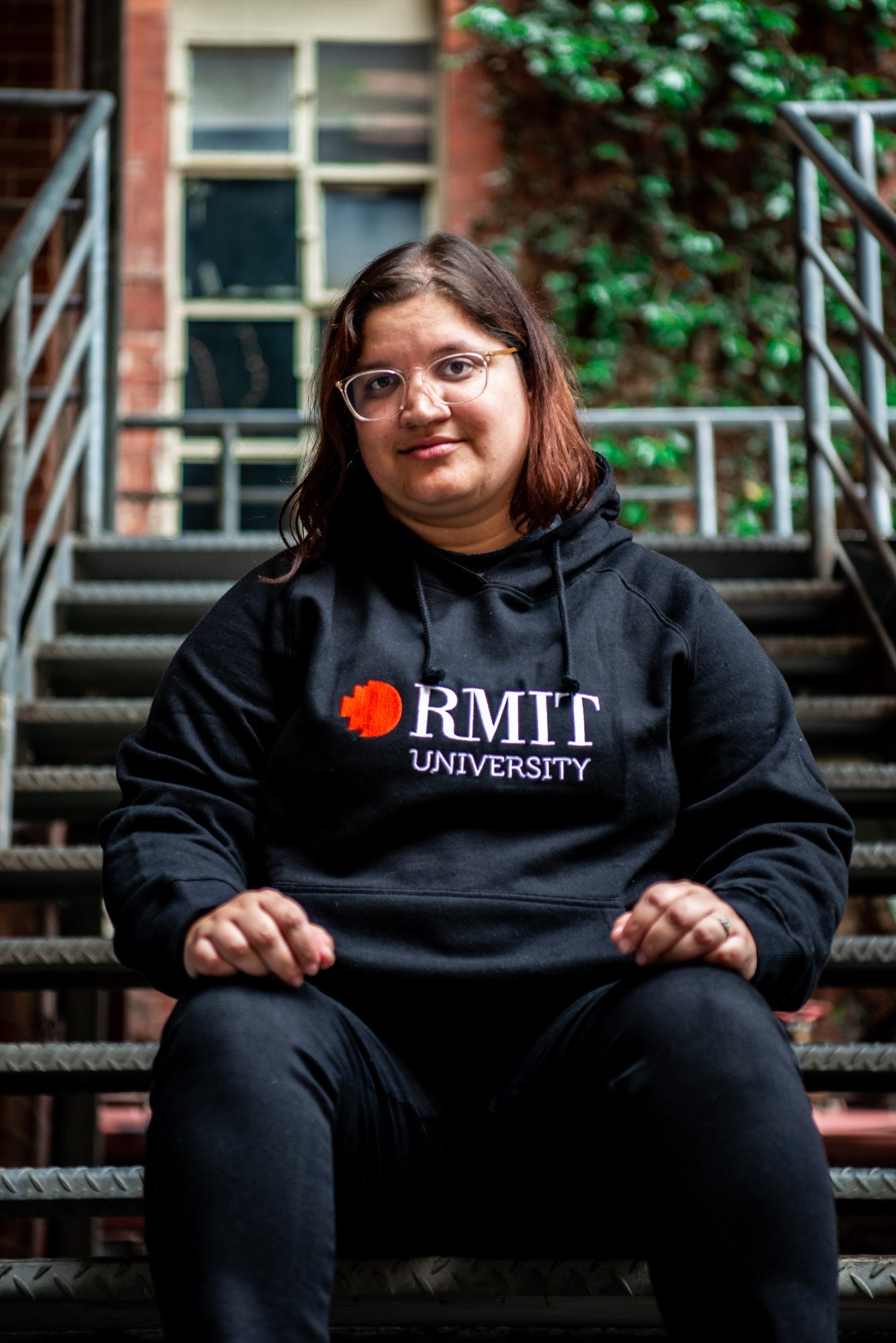 Female model wearing a black Fairtrade hoodie from the RMIT Store, facing forward with a relaxed pose. The hoodie features a red and white embroidered RMIT University logo centred on the chest. 