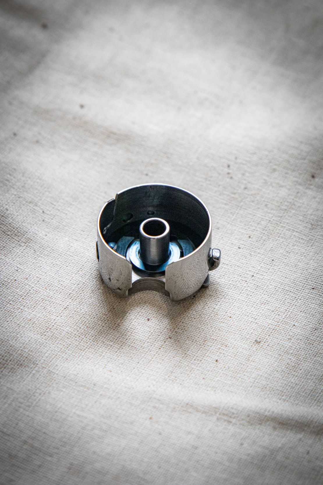 Bottom view of the RMIT Store bobbin case showing the central spindle and inner mechanism cavity, made of durable silver-toned metal, displayed on a lightly creased fabric background.