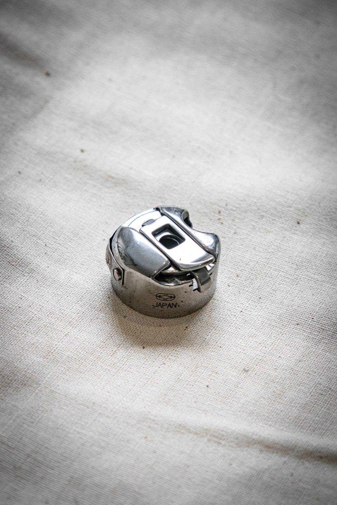 Close-up of a shiny silver RMIT Store bobbin case resting on a cream fabric surface, showing the top view with a rectangular opening and engraved "JAPAN" marking, made of polished metal for sewing machine compatibility.