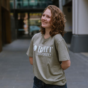 Model standing in an outdoor campus walkway wearing Bowen Tee in eucalyptus green featuring a large white RMIT University logo printed across the chest, styled with dark denim wide-leg jeans, smiling with hands behind back and head slightly turned, with blurred modern building background, available at the RMIT Store.