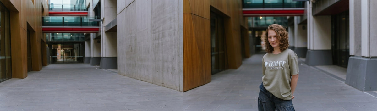 Model wearing the Bowen Tee in Eucalyptus, a short-sleeve crew-neck T-shirt with a large white RMIT University logo printed across the chest, styled with dark trousers and posed on the right side of a modern RMIT campus walkway with concrete walls, timber panelling and red architectural details for RMIT Store.