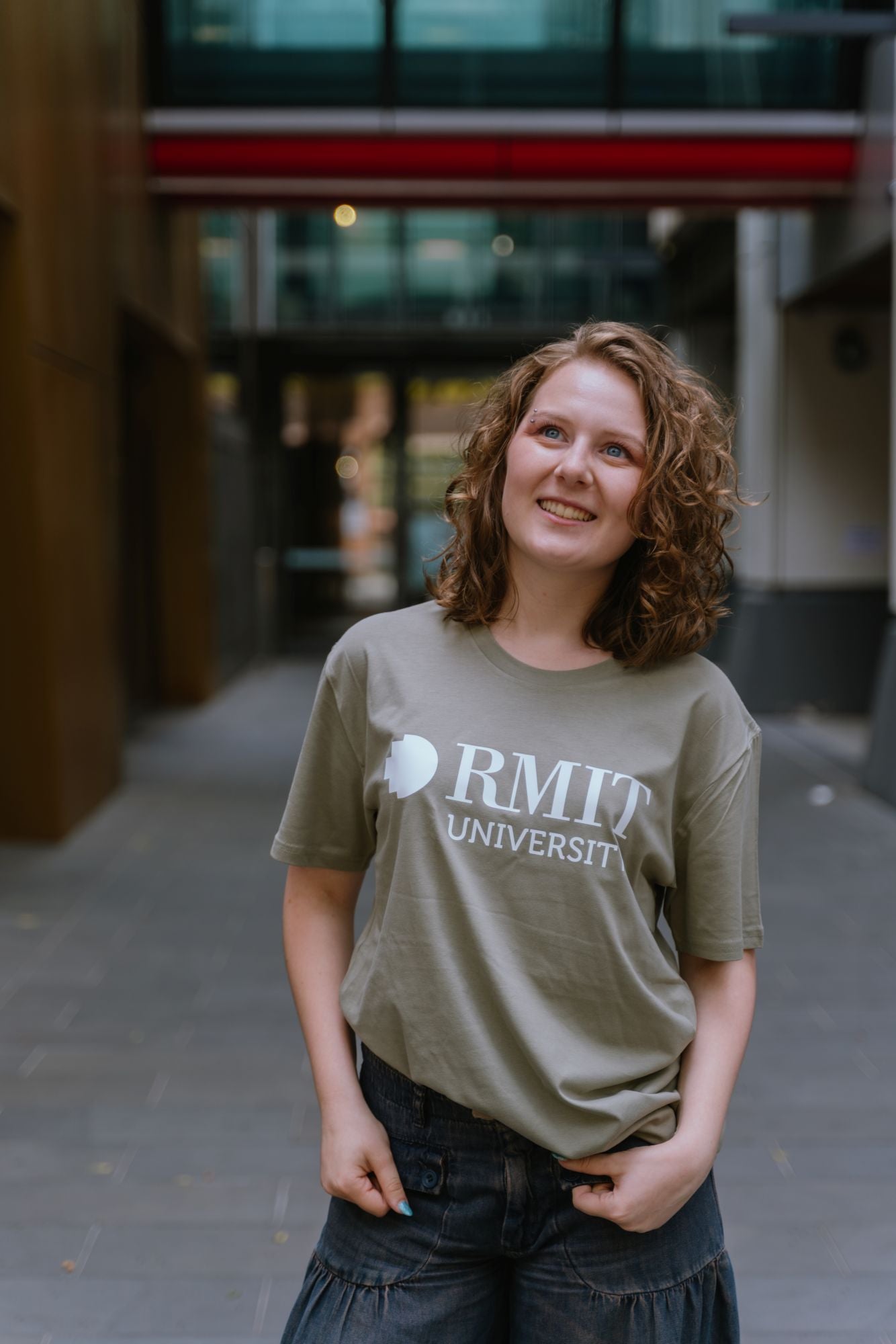 Model standing in an outdoor campus setting wearing Bowen Tee in eucalyptus green featuring large white RMIT University logo print across the chest, styled with dark denim wide-leg jeans, with one hand in pocket and relaxed stance against blurred architectural background, available at the RMIT Store.