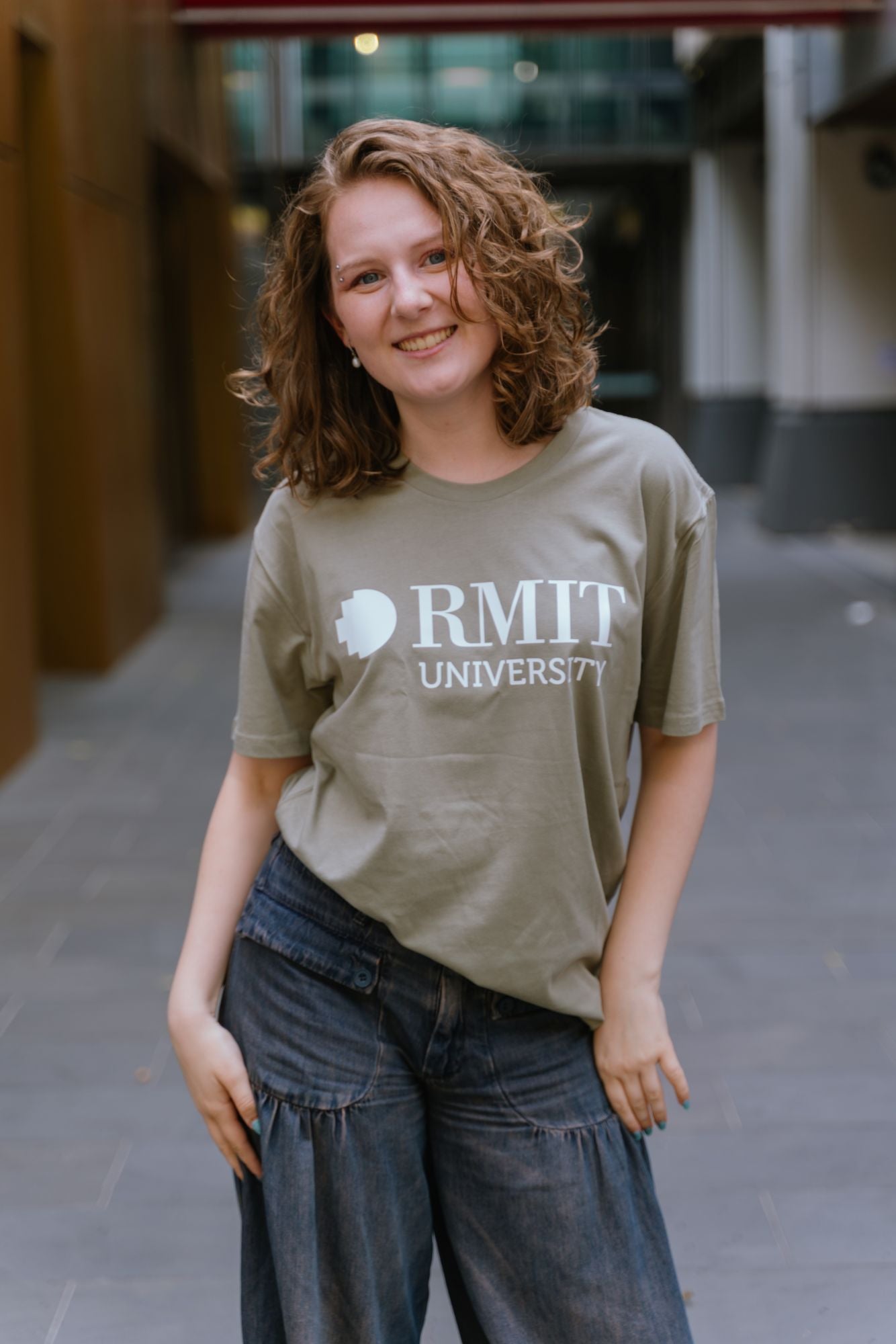 Model facing forward in an outdoor campus walkway wearing Bowen Tee in eucalyptus green with bold white RMIT University logo across the chest, paired with dark denim wide-leg jeans, smiling directly at camera with relaxed posture, available at the RMIT Store.