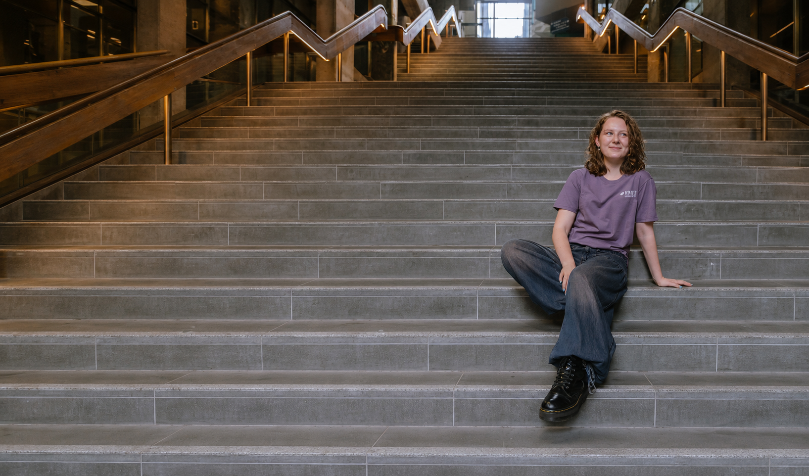 RMIT Store Bowen Tee in mauve, modelled from the front by a seated model with wavy brown hair, featuring a relaxed crew neck, short sleeves and one small white RMIT University logo printed on the left chest, styled with dark wide-leg jeans and black lace-up boots on a broad grey-tiled staircase with timber and glass handrails.