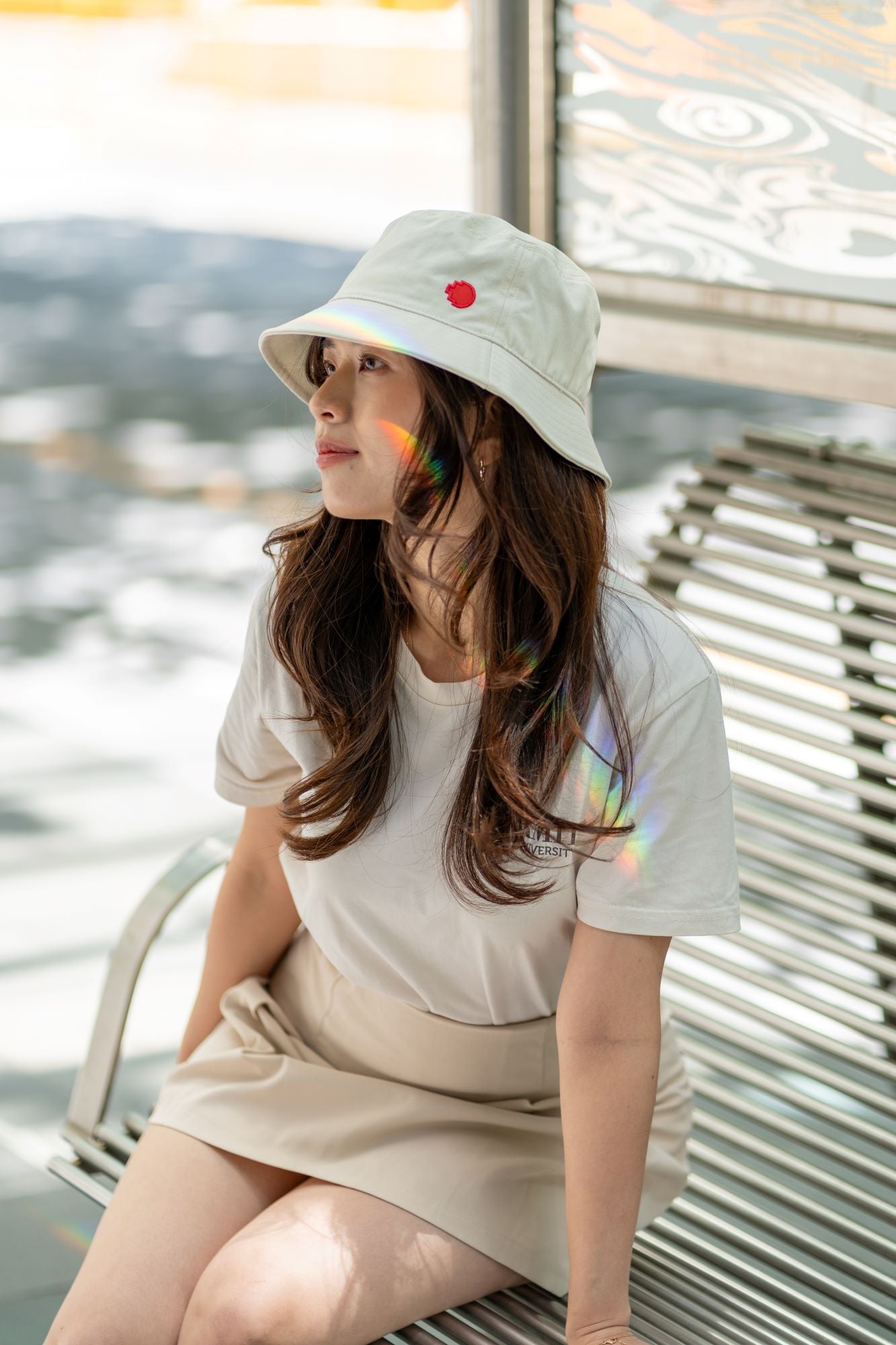 RMIT Store bucket hat in light beige, featuring the RMIT pixel logo, worn by a model in a beige skirt and white t-shirt, seated on a metallic bench. Rainbow light reflections add a dynamic touch to the chic urban casual outfit.