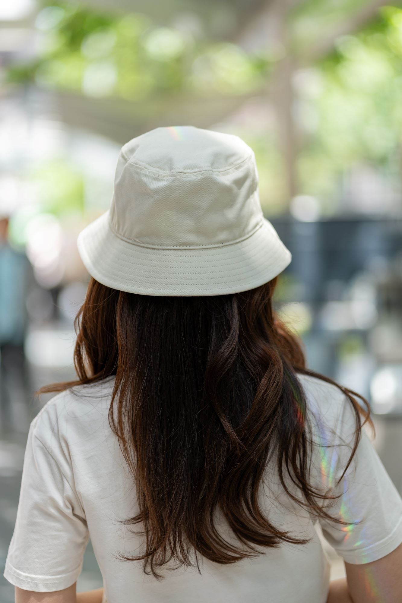 Light beige bucket hat from the RMIT Store, displayed from behind on a model with long, flowing hair. The blurred green background enhances the hat's relaxed fit and minimalist aesthetic.