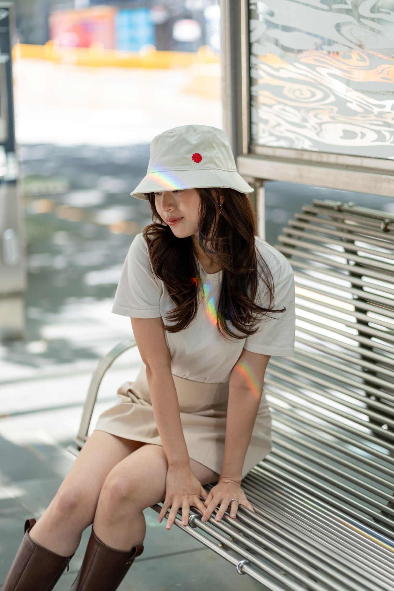 RMIT Store bucket hat in light beige with the RMIT pixel logo, paired with a beige skirt and white t-shirt. The model is seated on a bench in an outdoor setting, capturing a stylish urban look with soft lighting.