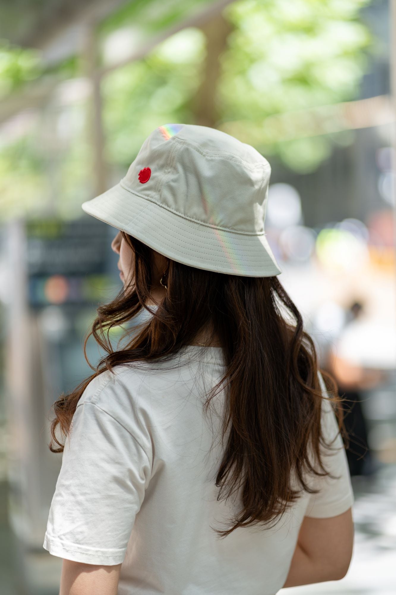 RMIT Store bucket hat in light beige, featuring the RMIT pixel logo in red on the side. The model's long hair complements the white t-shirt, set against a backdrop of lush green foliage, highlighting the hat's casual urban style.