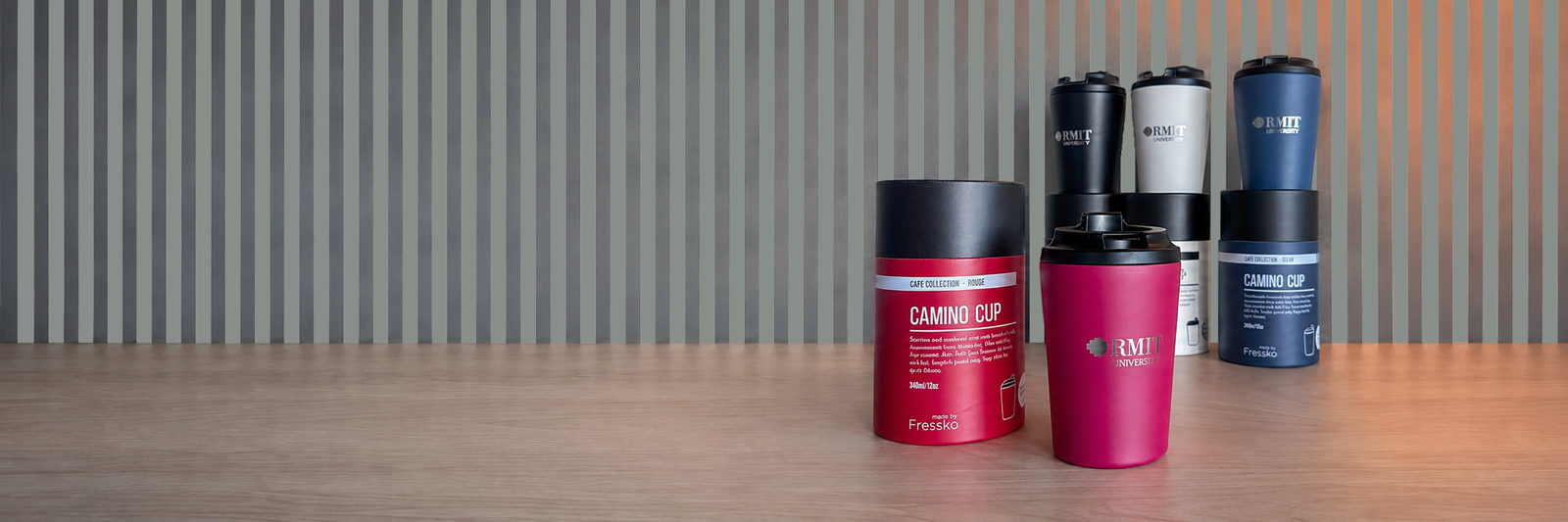 Four RMIT Store Camino Cups in Rouge, Frost and Denim, Coal, displayed with black lids and silver RMIT University logos alongside four matching cylindrical Fressko packages on a timber table, against a grey-and-white striped wall with glowing amber glass lights.
