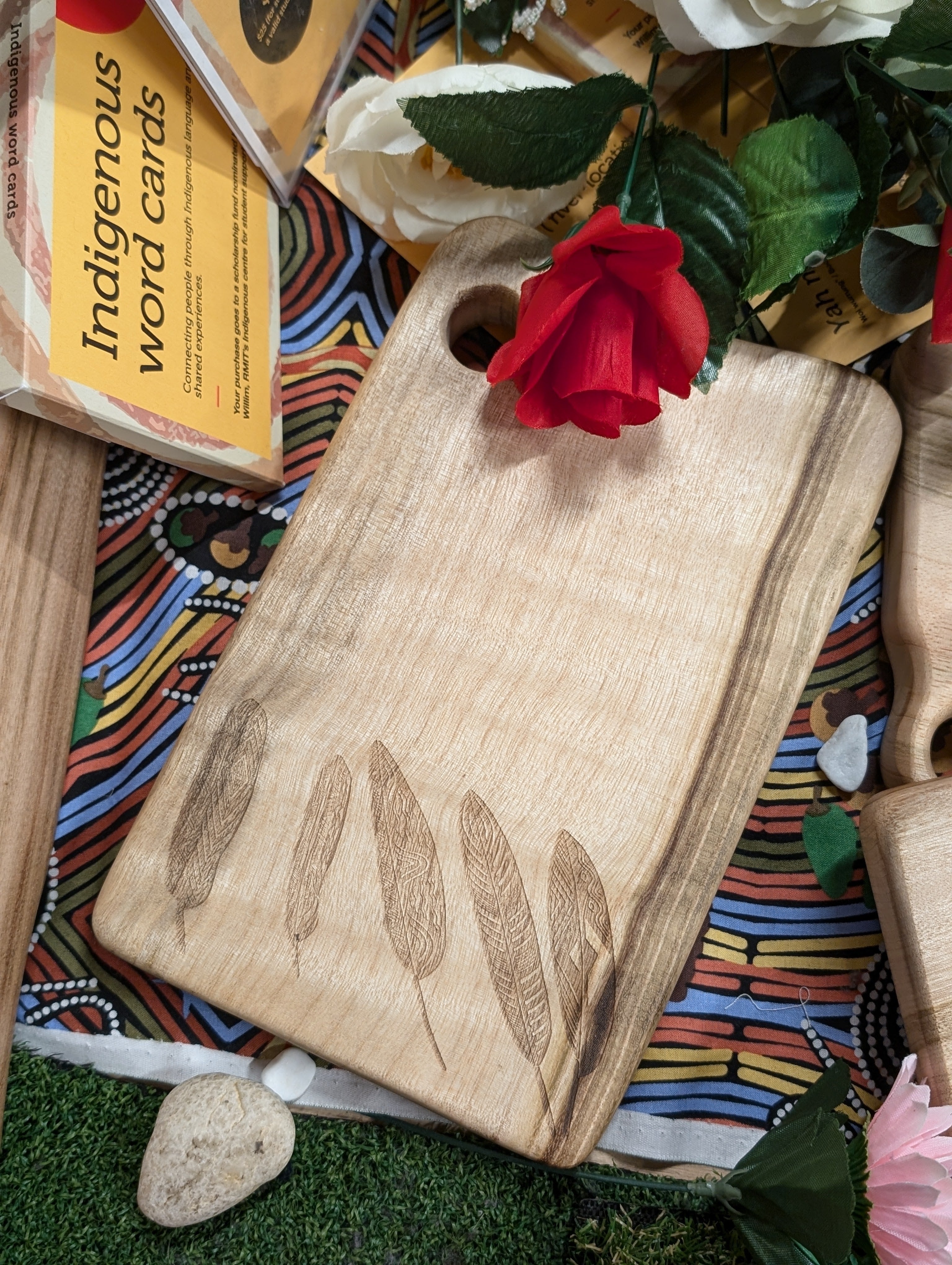An RMIT Store Camphor Laurel Board Mini Rectangle resting on a patterned cloth, surrounded by flowers and Indigenous word cards. The board displays engraved feather designs, highlighting detailed craftsmanship and the natural wood grain, set in a vibrant cultural context.