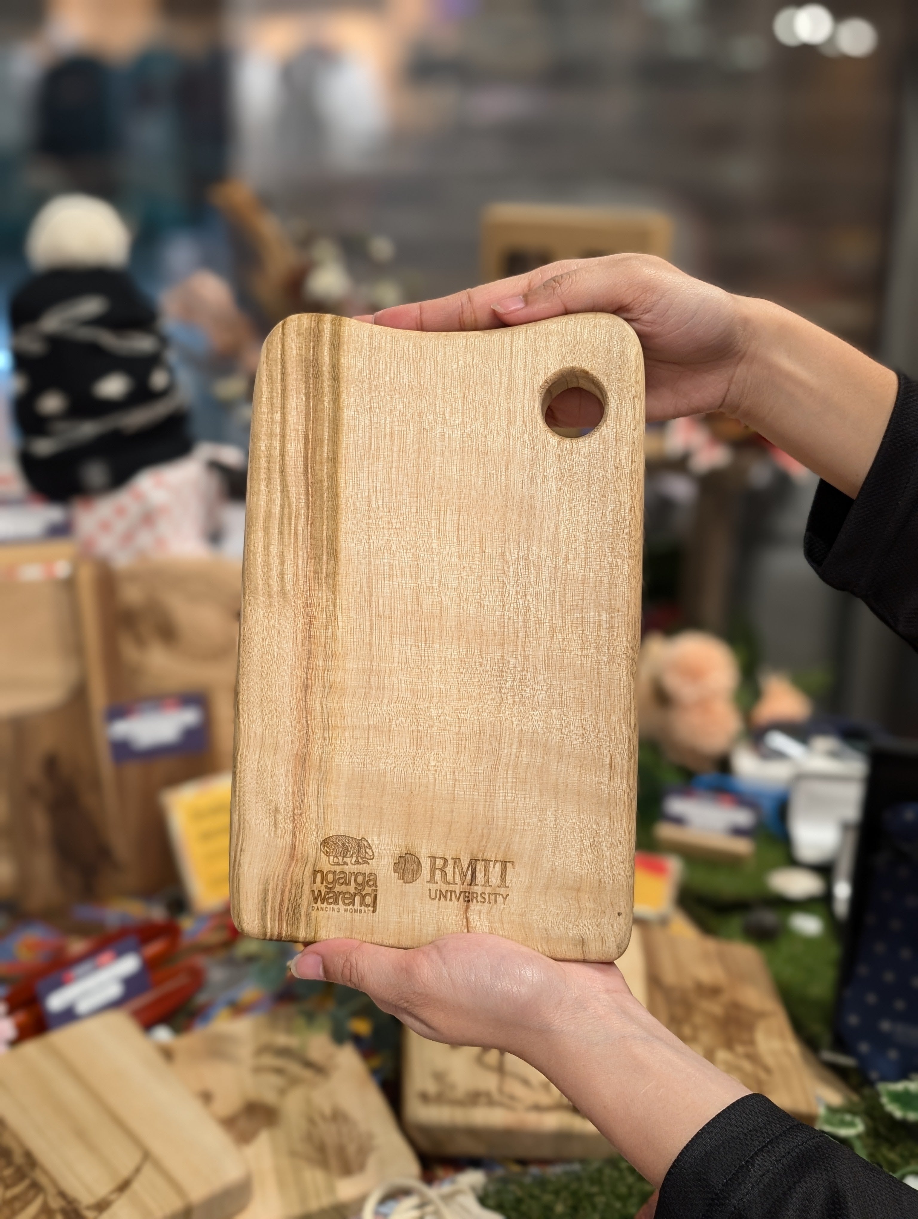 Hands holding an RMIT Store Camphor Laurel Board Mini Rectangle, showcasing its smooth wooden surface with intricate grain patterns at the back. The board features a circular cut-out for easy hanging and engraved logos of Ngarga Warendj Dancing Wombat and RMIT University, adding a touch of authenticity and branding.