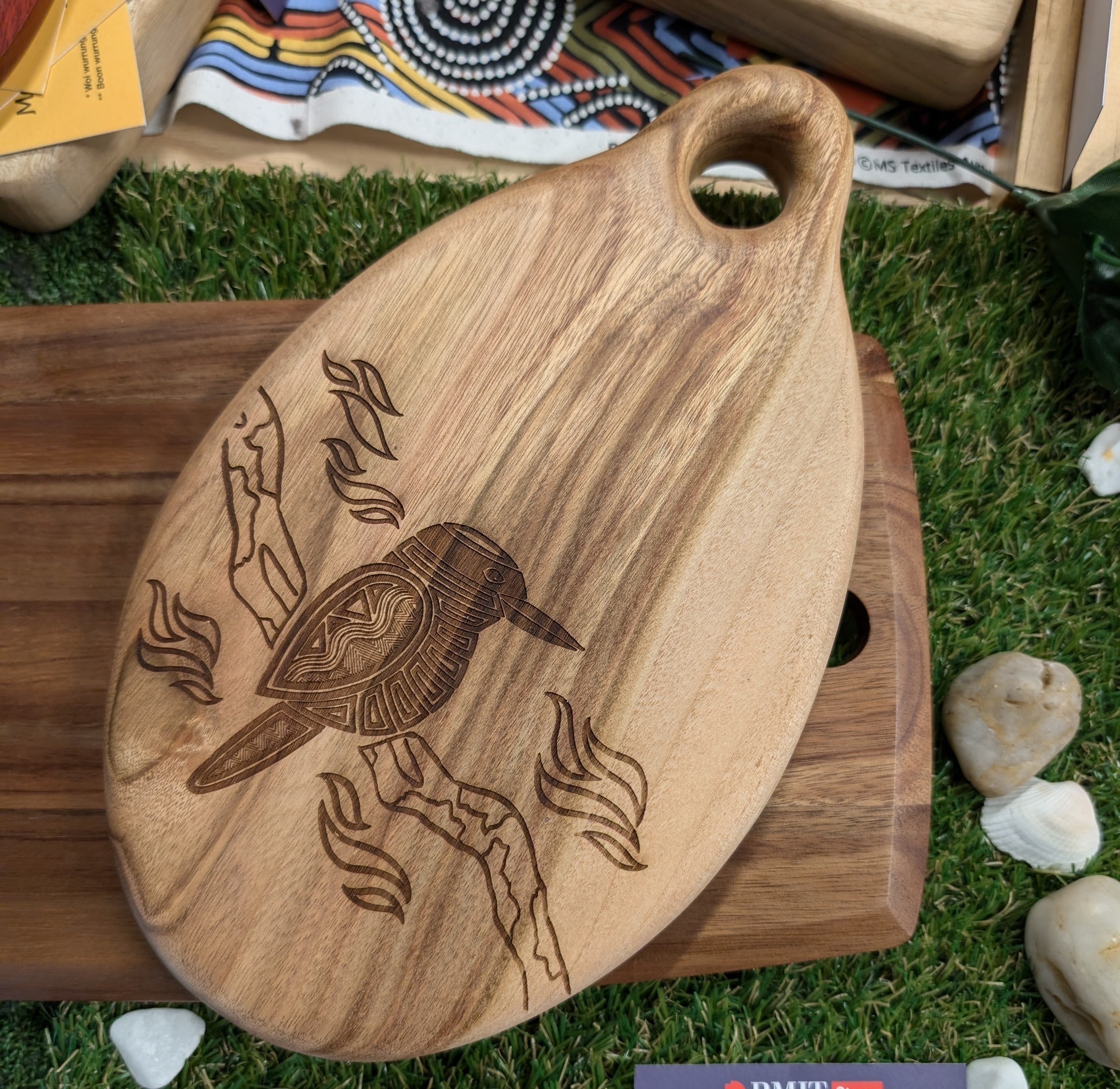 RMIT Store Camphor Laurel Board Oval Small placed on green turf. The board features a detailed engraved kookaburra design, highlighting the unique wood grain patterns and the board's curved shape with a handle cut-out for easy handling.
