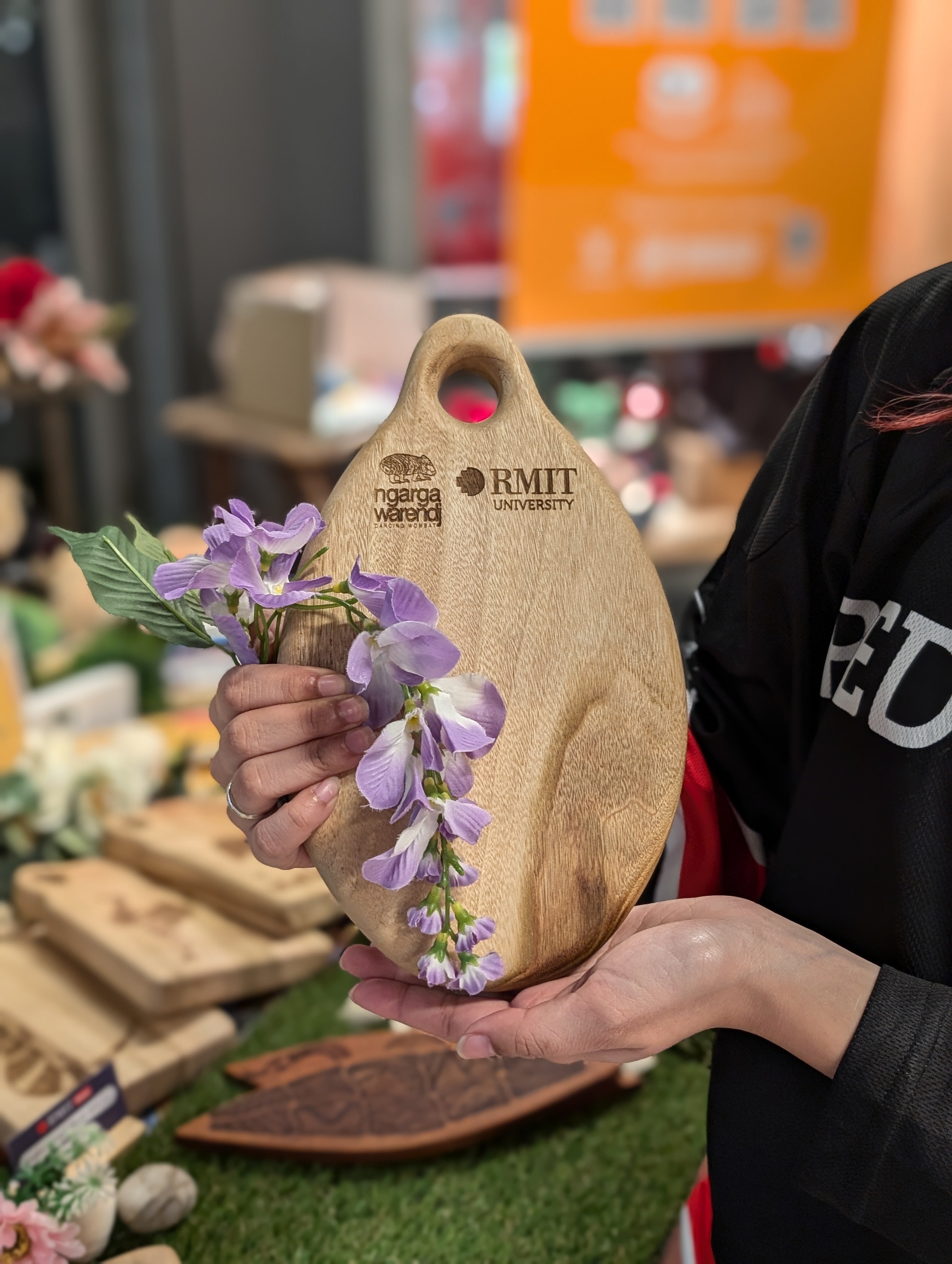 Hands holding an RMIT Store Camphor Laurel Board Oval Small, adorned with a cascade of vibrant purple flowers. The smooth wooden surface displays engraved logos of Ngarga Warendj Dancing Wombat and RMIT University, reflecting craftsmanship and branding.
