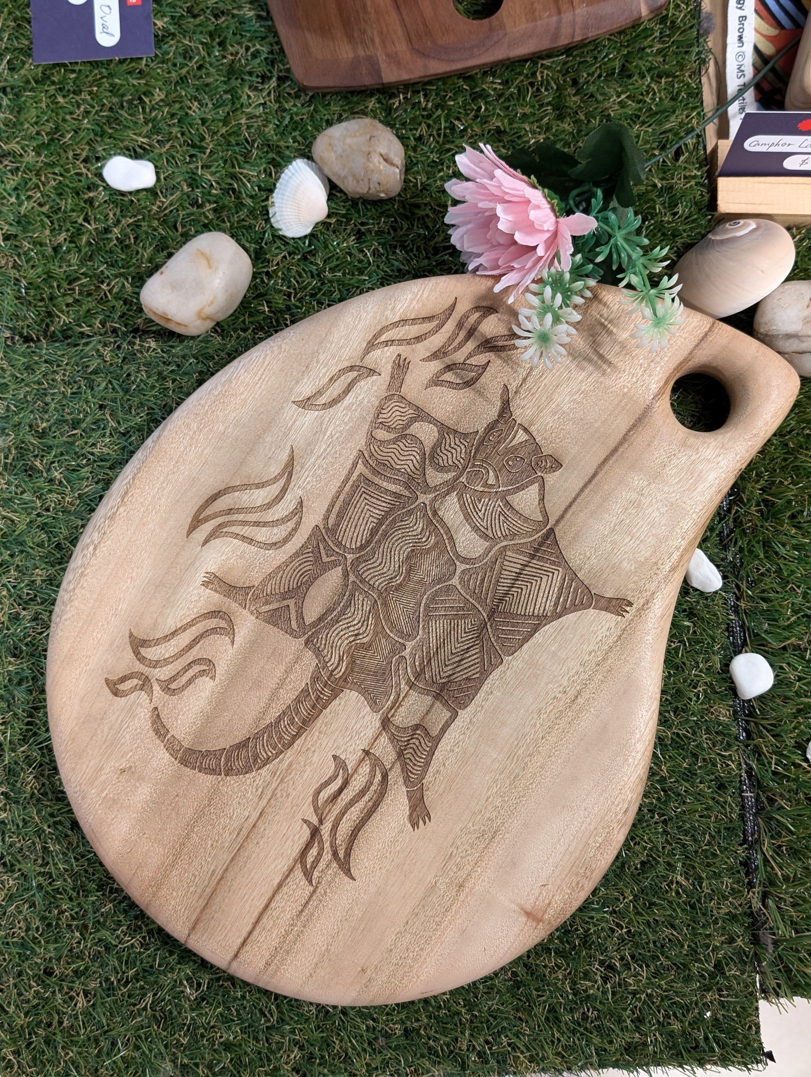 RMIT Store Camphor Laurel Board Tear displayed on grass, adorned with engraved art of a possum glider surrounded by intricate patterns. Flowers and rocks accent the setting, highlighting the finely detailed craftsmanship and unique tear-shaped board design.