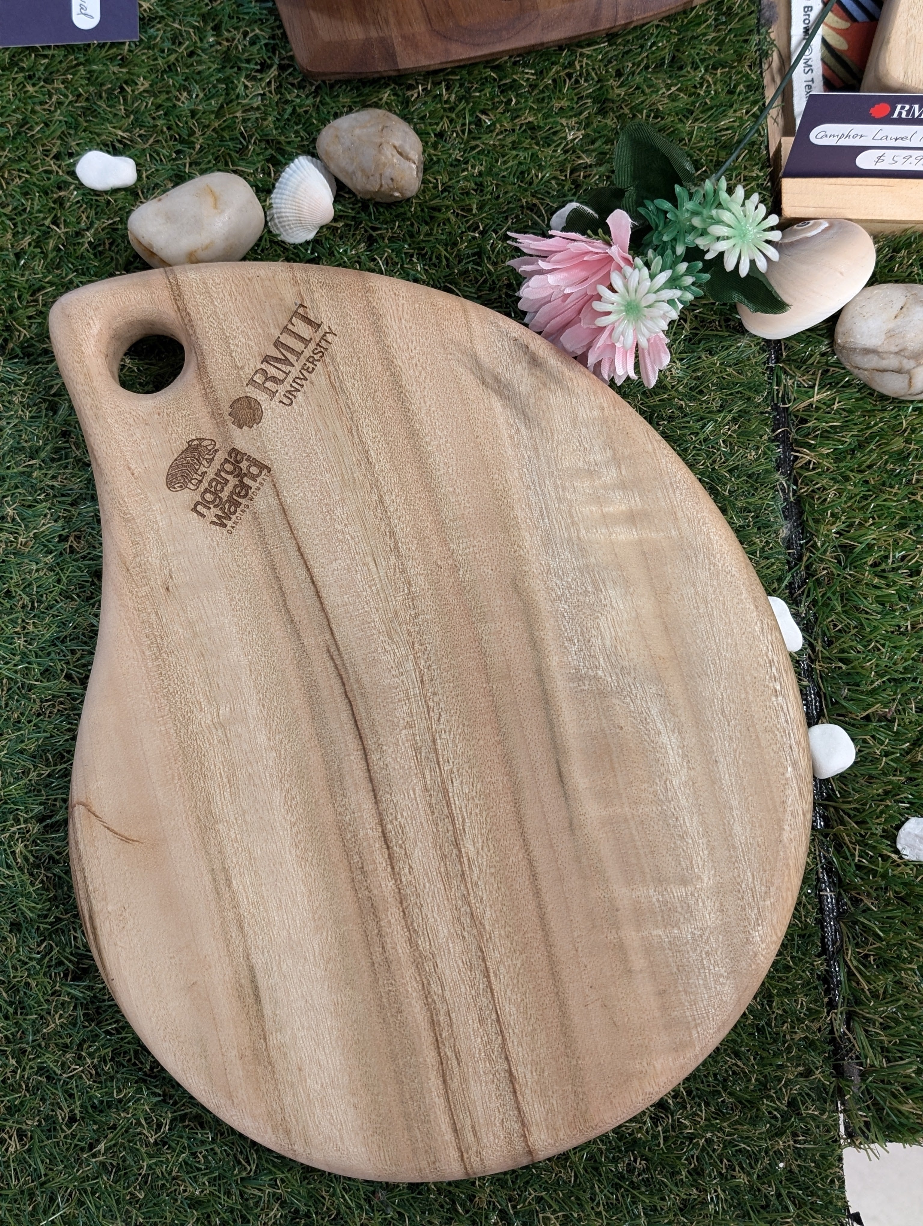 RMIT Store Camphor Laurel Board Tear placed on a grass surface with scattered rocks and flowers. The board features a tear-shaped design with smooth, natural wood grain patterns and a convenient handle cut-out. Engraved logos of Ngarga Warendj Dancing Wombat and RMIT University are visible, emphasizing craftsmanship and branding.