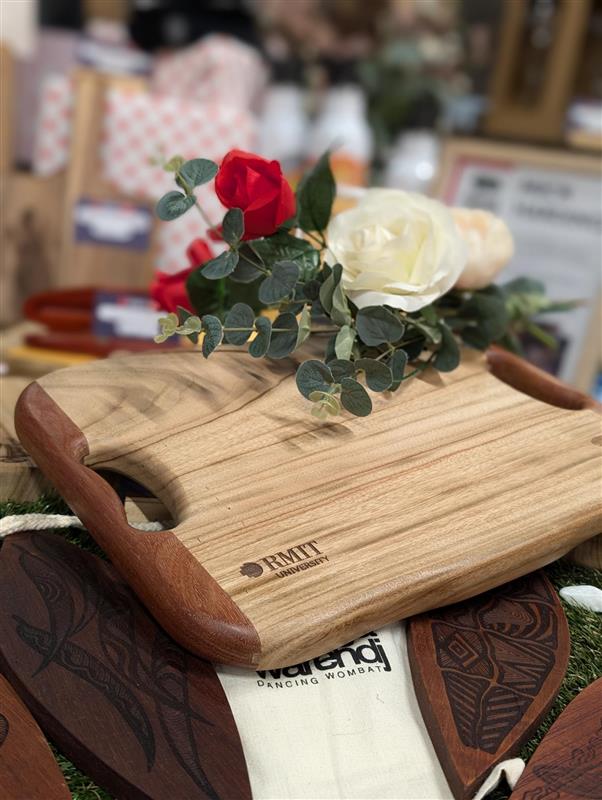 Camphor Laurel and Redgum board with handle from the RMIT Store, engraved with RMIT University branding. It features a mix of light and dark wood tones, styled with red and white floral decorations, and is displayed on a wooden surface.
