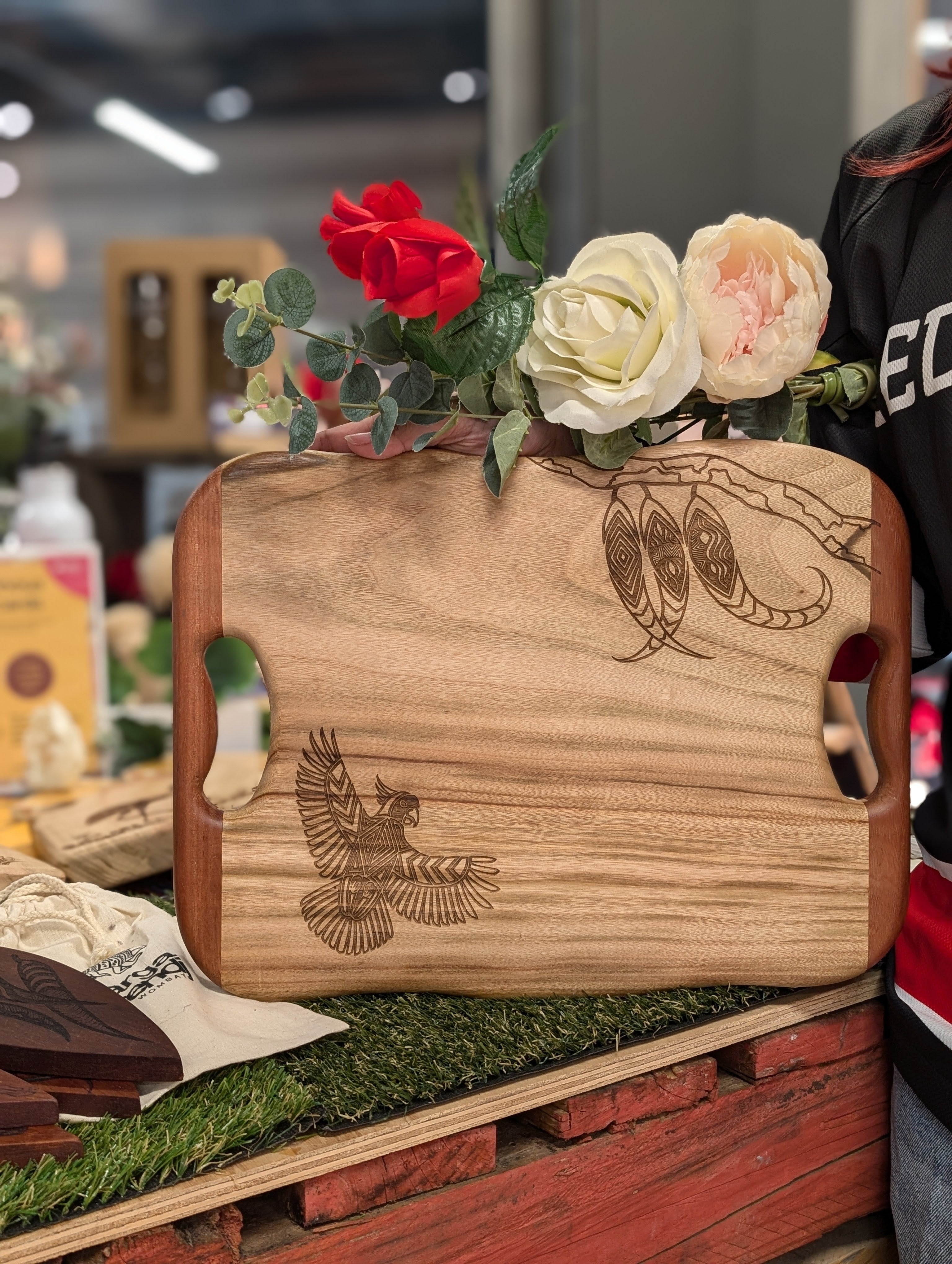 Detailed view of the Camphor Laurel and Redgum board from the RMIT Store, highlighting an intricate engraving of a cockatoo. The board, held by a person wearing a dark shirt, is adorned with red, white, and pink roses, illustrating its elegant design.