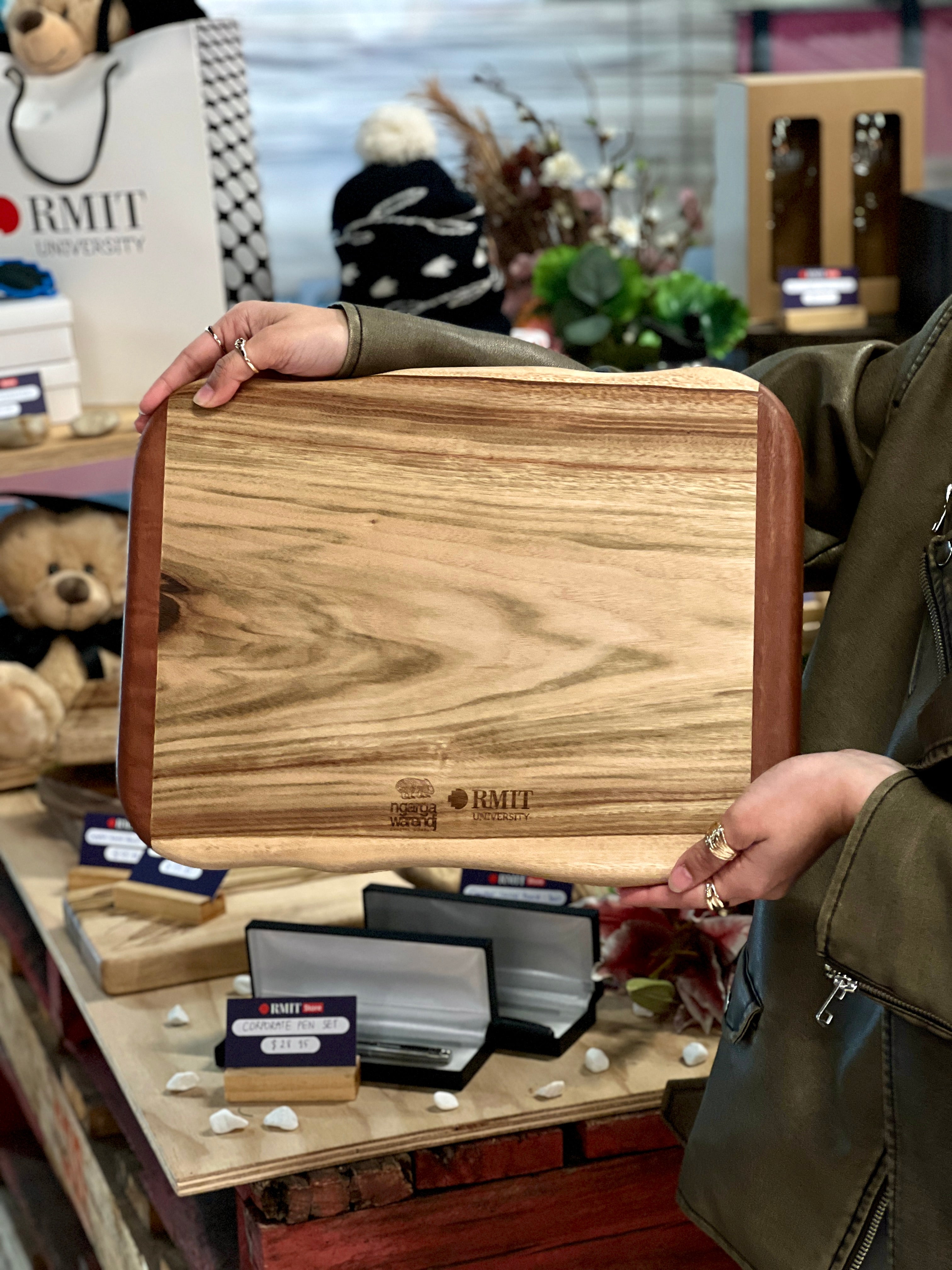 Camphor Laurel and Redgum board held by a model, featuring a smooth natural grain pattern and RMIT University branding, displayed among RMIT Store products including gift bags and accessories.