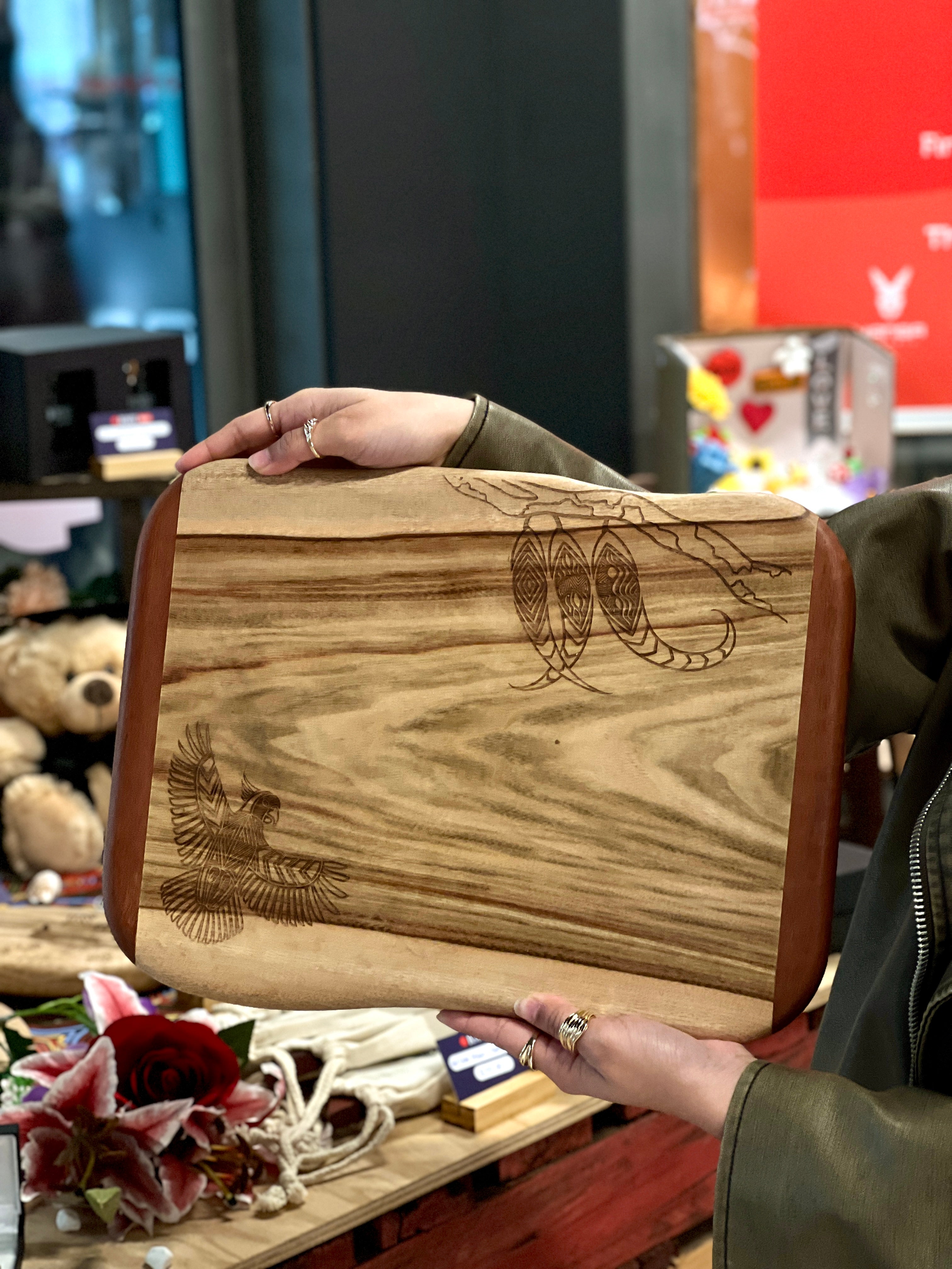 Camphor Laurel and Redgum board with indigenous artwork engravings including a cockatoo and intricate patterns, held by a model in the vibrant setting of the RMIT Store, surrounded by a graduation bear and items from the same collection.