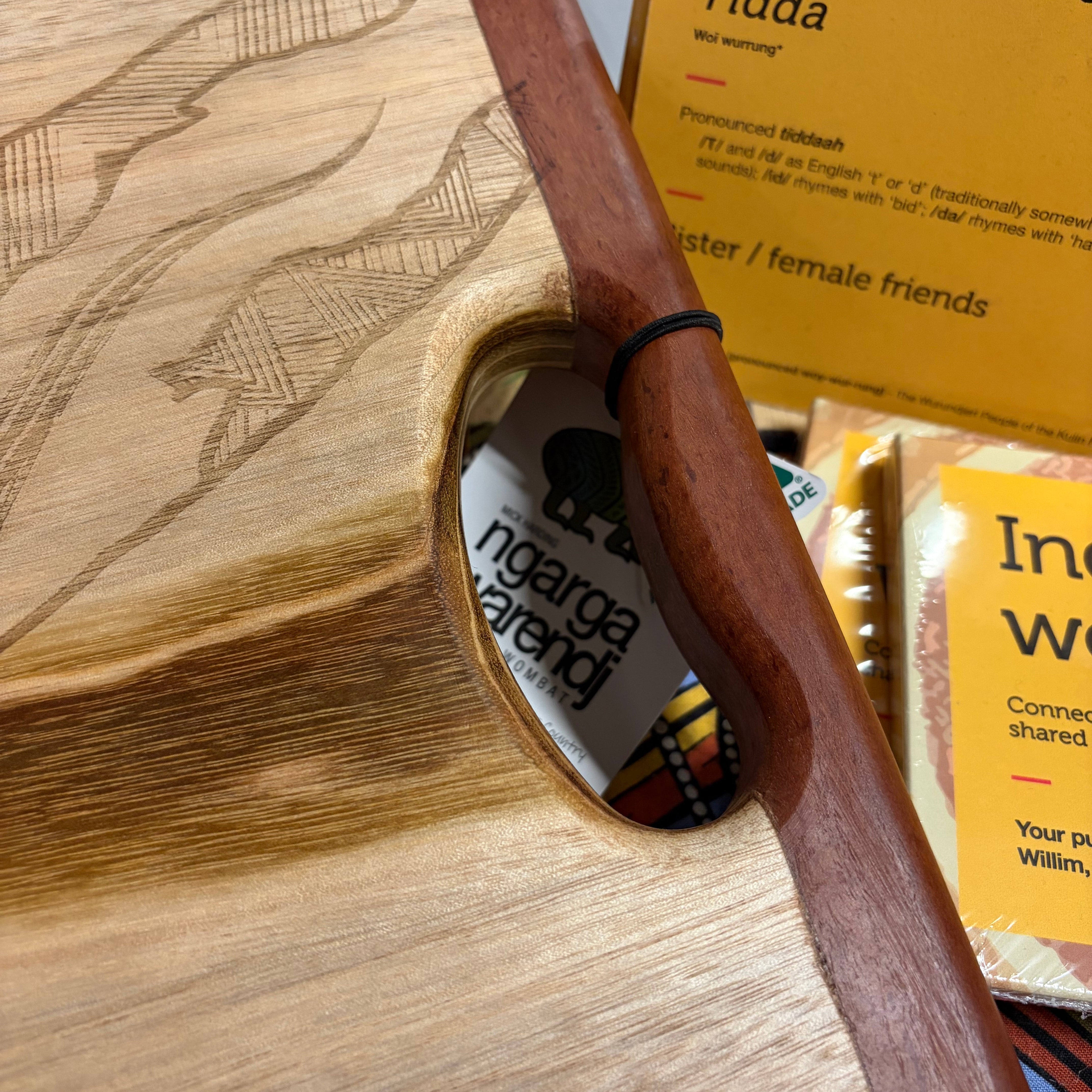 Close-up of Camphor Laurel & Redgum Board with handle focusing on the smooth curved redgum handle and detailed laser-etched Dhulin the Goanna artwork on the camphor laurel surface, showing fine linework patterns and natural wood grain variations; positioned beside yellow Indigenous language cards and store display items, photographed on a light grey surface at the RMIT Store.