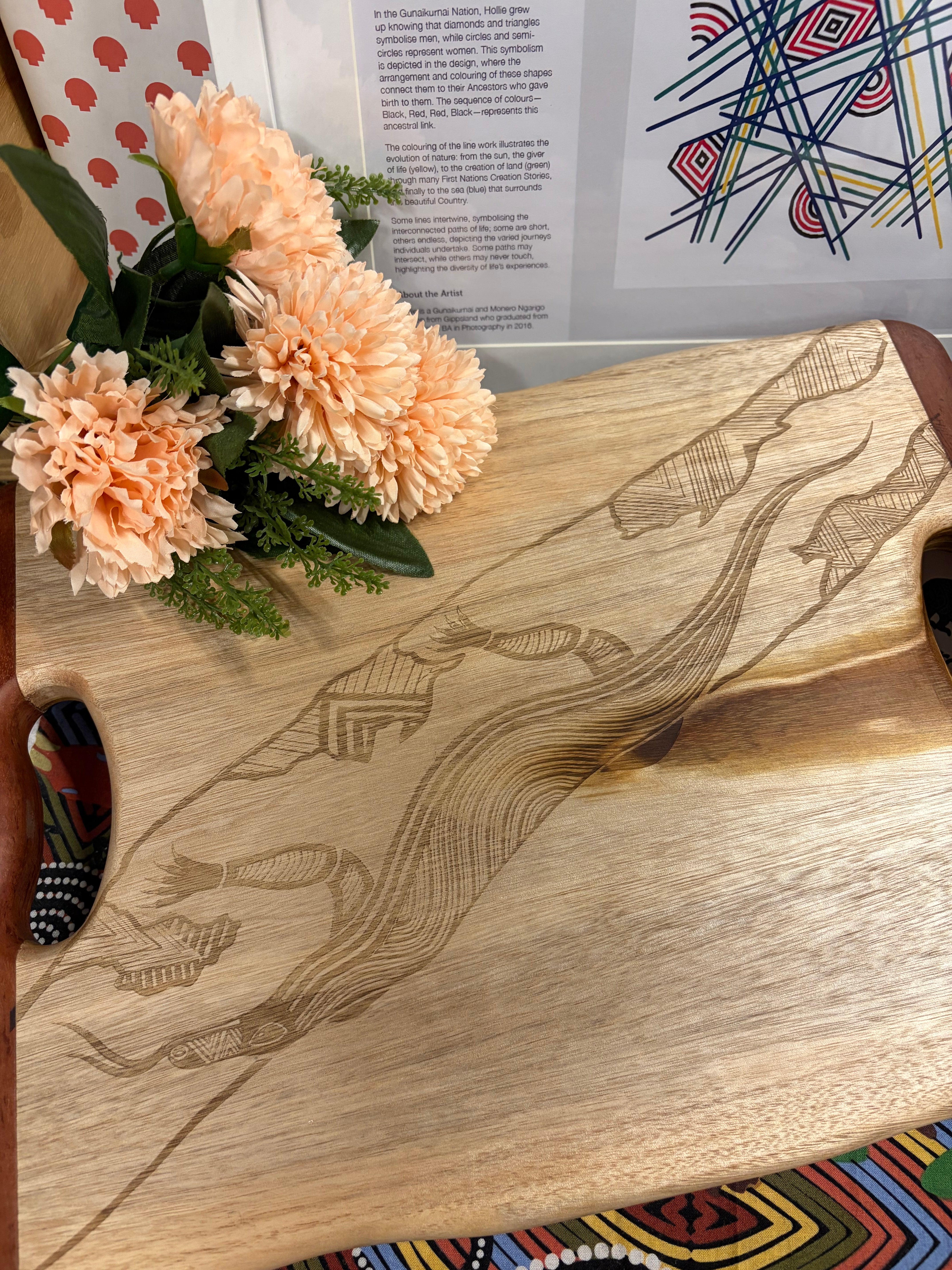 Camphor Laurel & Redgum Board with handle styled in an in-store display at the RMIT Store, showing full board with rich natural timber grain, contrasting redgum handles, and intricate laser-etched Dhulin the Goanna design spanning the surface; surrounded by Indigenous-inspired merchandise, floral arrangement, and framed artwork, highlighting cultural context and retail presentation.