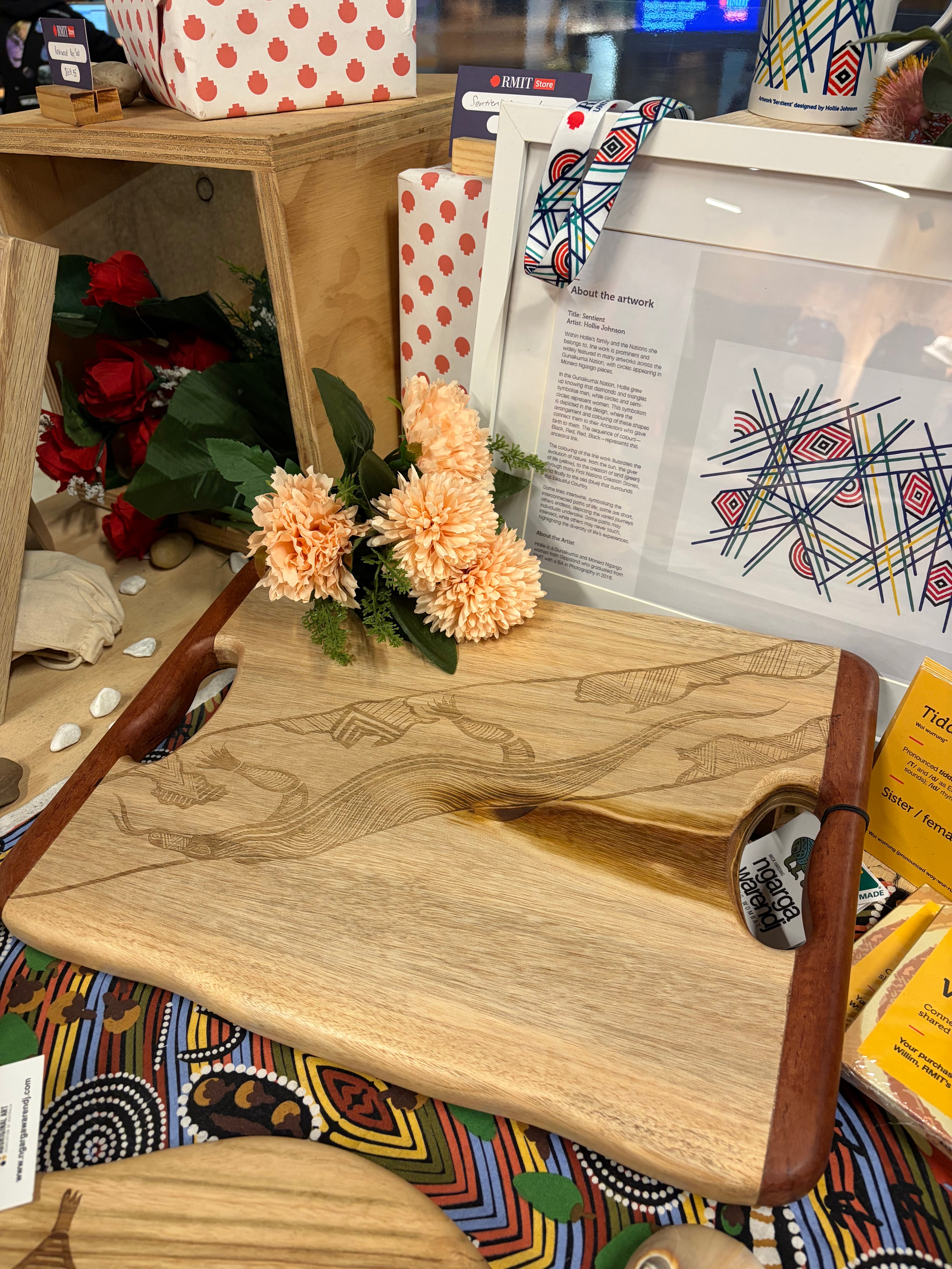 Camphor Laurel & Redgum Board with handle displayed on a light grey surface, featuring a natural camphor laurel wood grain with darker redgum handles at each end, and a detailed laser-etched Dhulin the Goanna artwork with multiple traditional Aboriginal design elements including line patterns and animal motifs across the centre; styled with peach artificial flowers and cultural artwork prints in the background, photographed in-store at the RMIT Store.