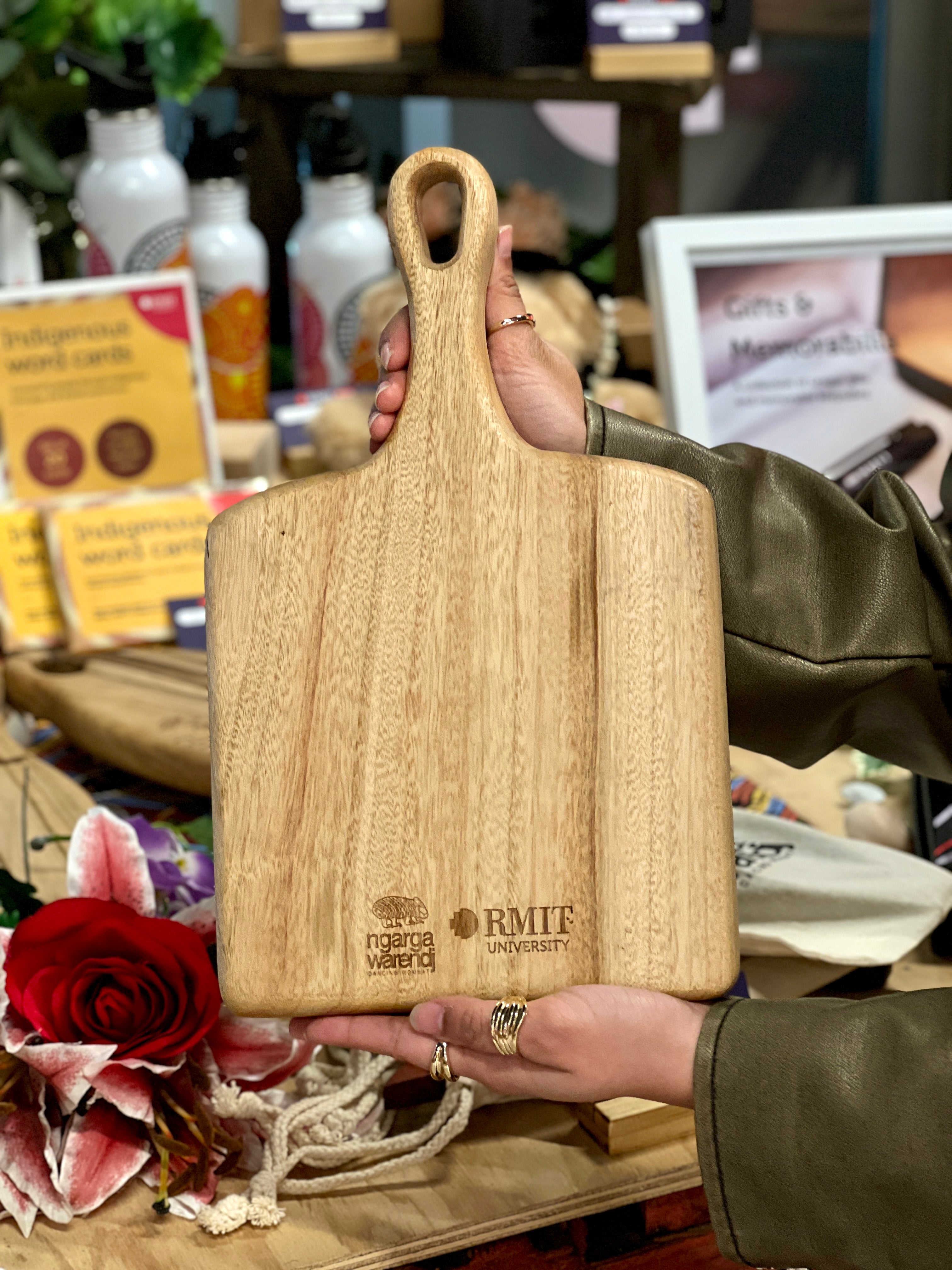 Back view of RMIT Store Camphor Laurel Square Paddle showcasing the engraved RMIT University and Ngarga Warendj logos. The paddle is held against a backdrop of stylish Indigenous-themed products.