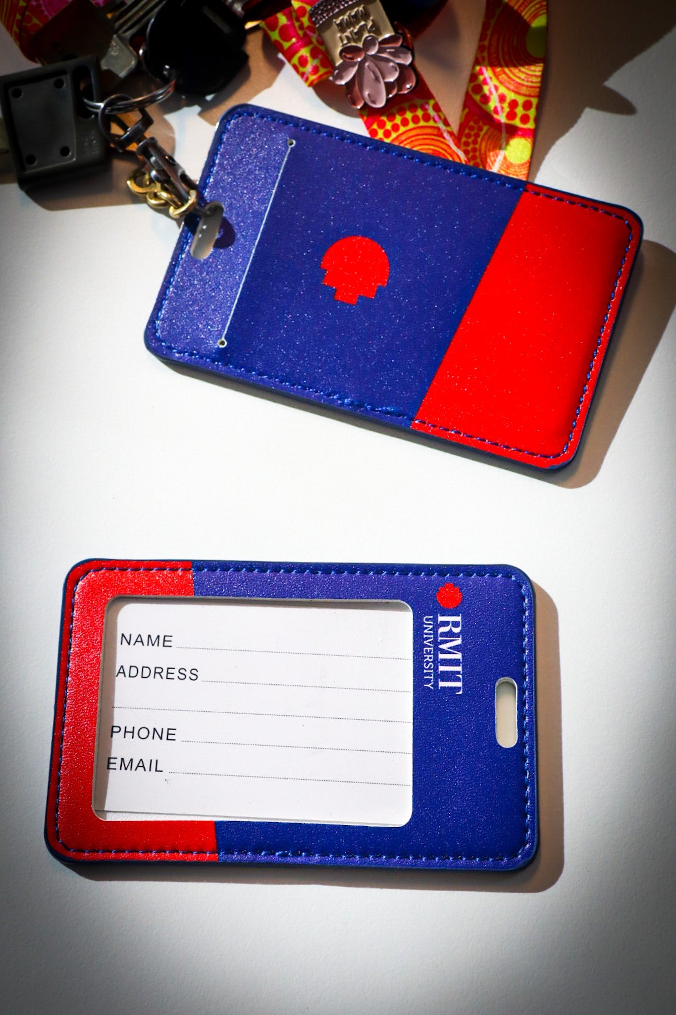 Close-up view of two RMIT Store cardholders on a white surface. The top holder, attached to a vibrant patterned lanyard with keys, shows a striking blue and red design, complete with the RMIT logo. The lower holder highlights fields for name, address, phone, and email, emphasising its utility for identification.
SEO File Names