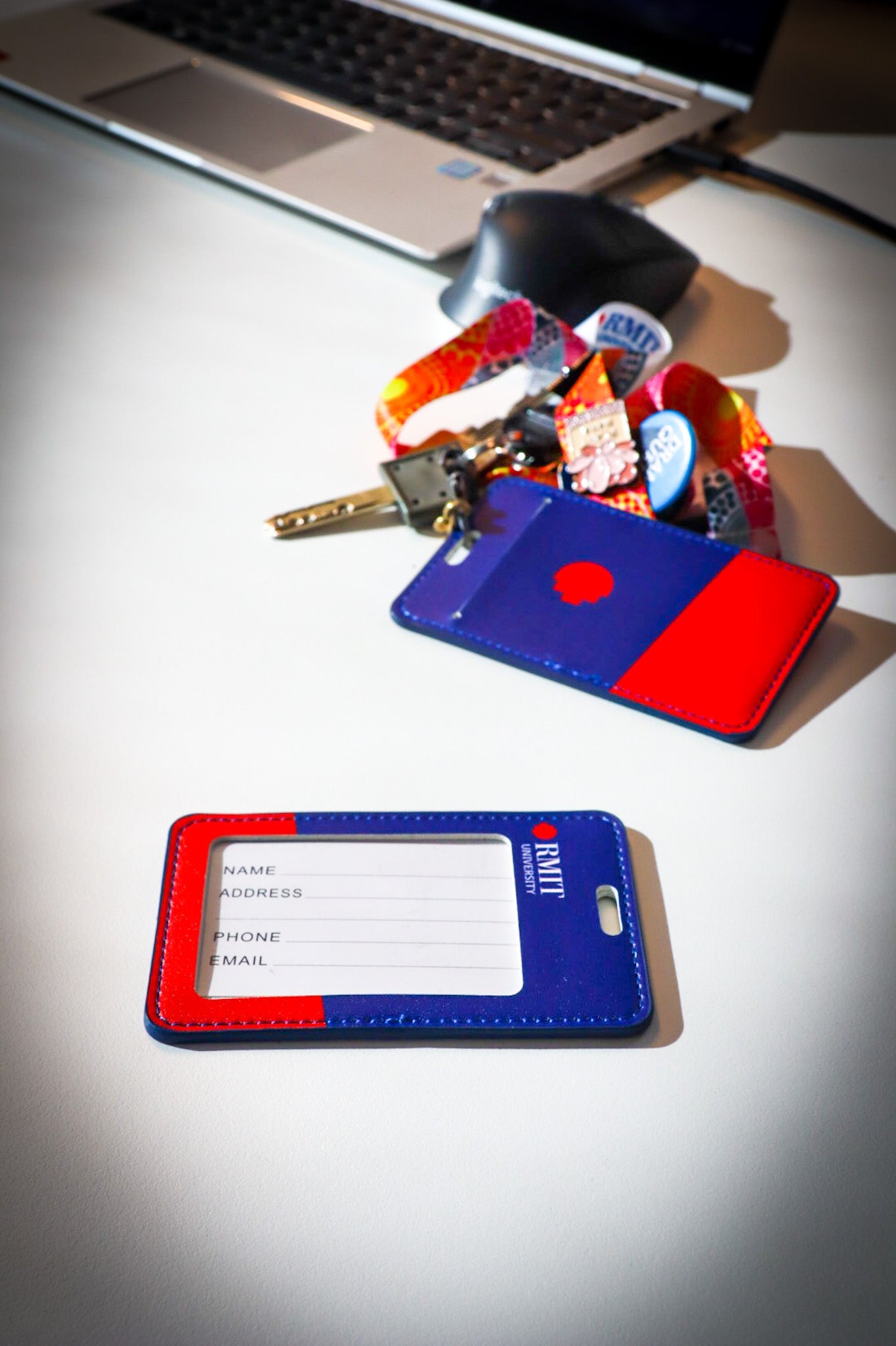 Two cardholders from the RMIT Store sit on a white desk beside a laptop. The lower card holder is own. The upper one is attached to a vibrant lanyard with keys and pins.