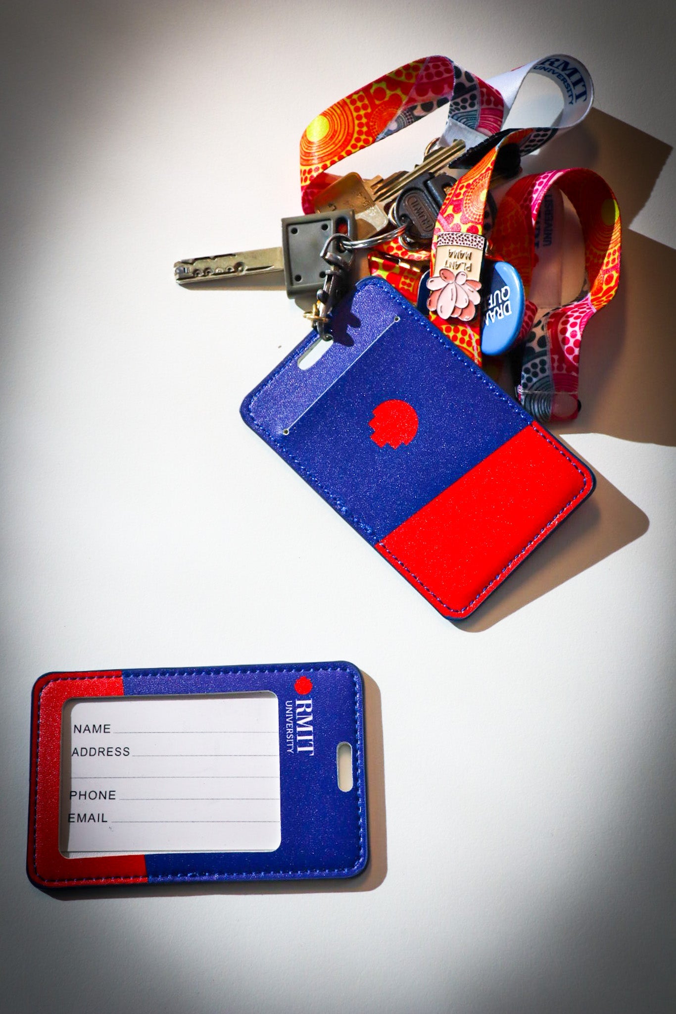 Two RMIT Store cardholders are displayed on a white surface. The cardholders are blue and red, featuring the RMIT logo. One is attached to a colourful lanyard with keys and decorative pins and badges, while the other shows spaces for personal information.
