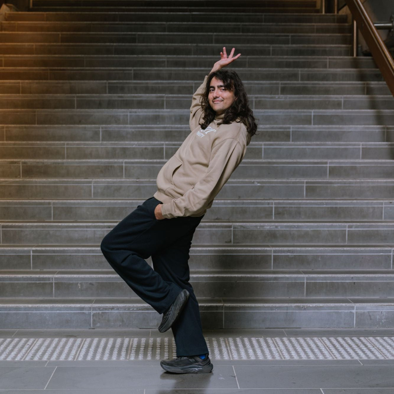 Model posing playfully on indoor staircase wearing Casey Hoodie in sand with bold white screen-printed RMIT University logo across the chest, one hand raised and one in pocket, paired with black pants in a modern architectural stairwell, available at the RMIT Store.
