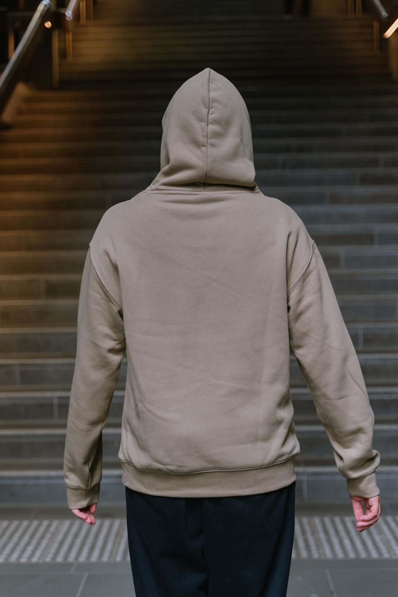 Rear view of model standing on indoor staircase wearing RMIT Casey Hoodie in sand, featuring hood up with plain back and no visible print, relaxed fit with ribbed hem and cuffs, styled with black pants in a modern stairwell setting, available at the RMIT Store.