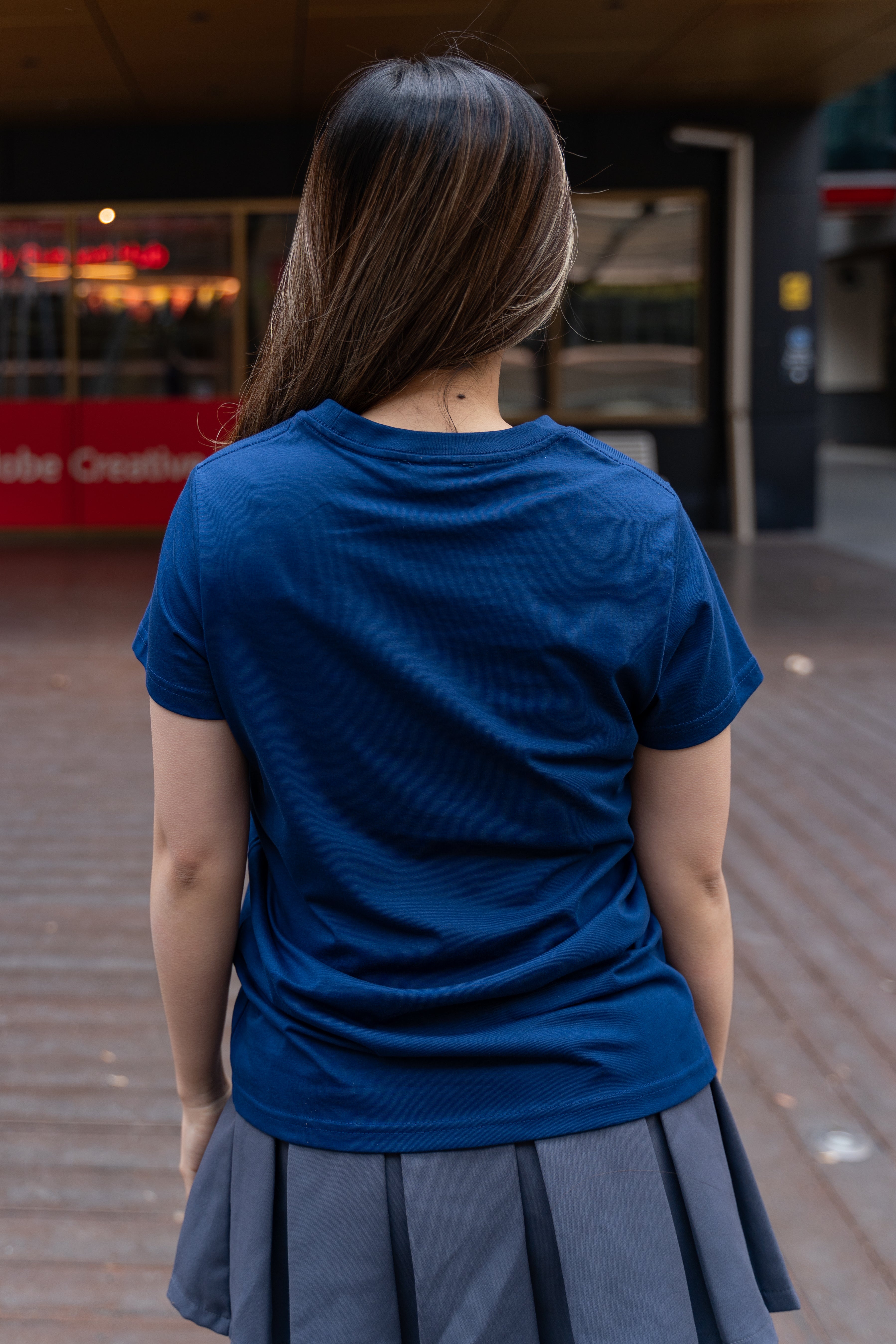 Female model shown from the back wearing a cobalt blue RMIT Class of 2025 Tee with plain rear design, short sleeves, and round neckline, styled with a pleated grey skirt in an outdoor campus walkway, available at the RMIT Store.