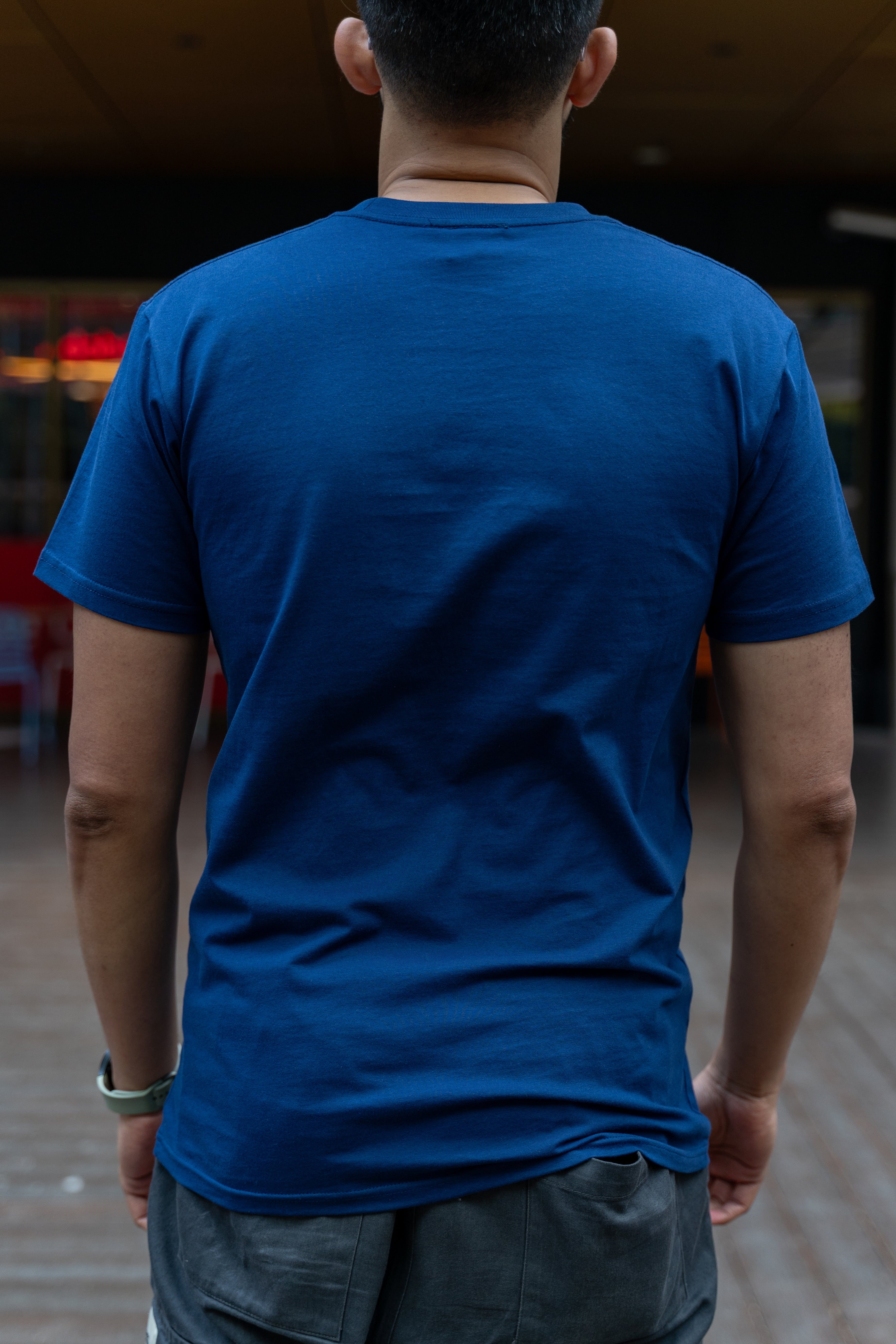 Male model shown from behind wearing a cobalt blue RMIT Class of 2025 Tee with a plain back, short sleeves, and straight hem, standing on an outdoor campus walkway, available at the RMIT Store.