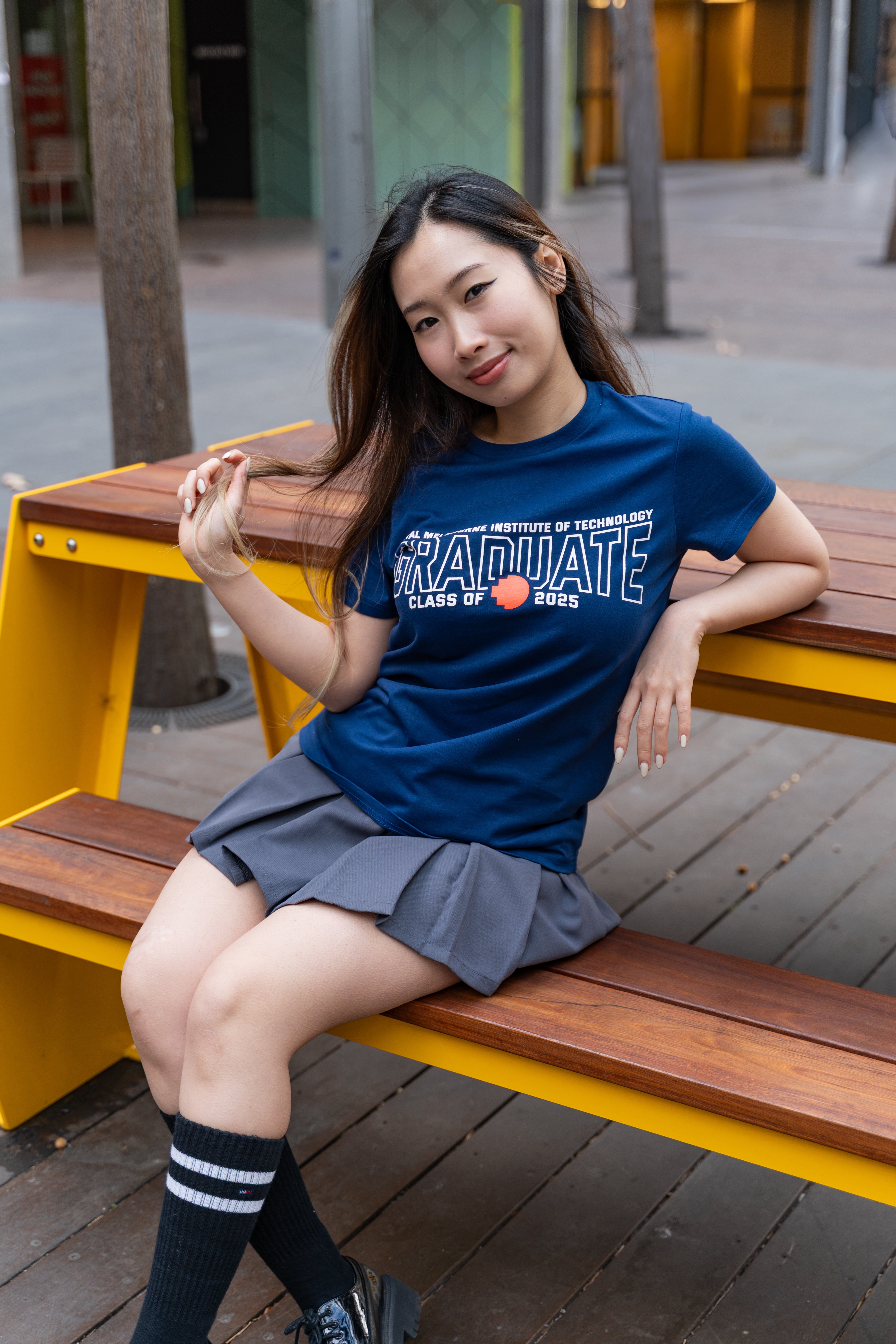 Female model wearing a cobalt blue RMIT Class of 2025 Tee with bold white “Graduate” text across the chest, featuring the red pixel RMIT logo; styled with a pleated grey skirt and black striped knee socks while seated on an outdoor wooden bench with yellow frame, available at the RMIT Store.
