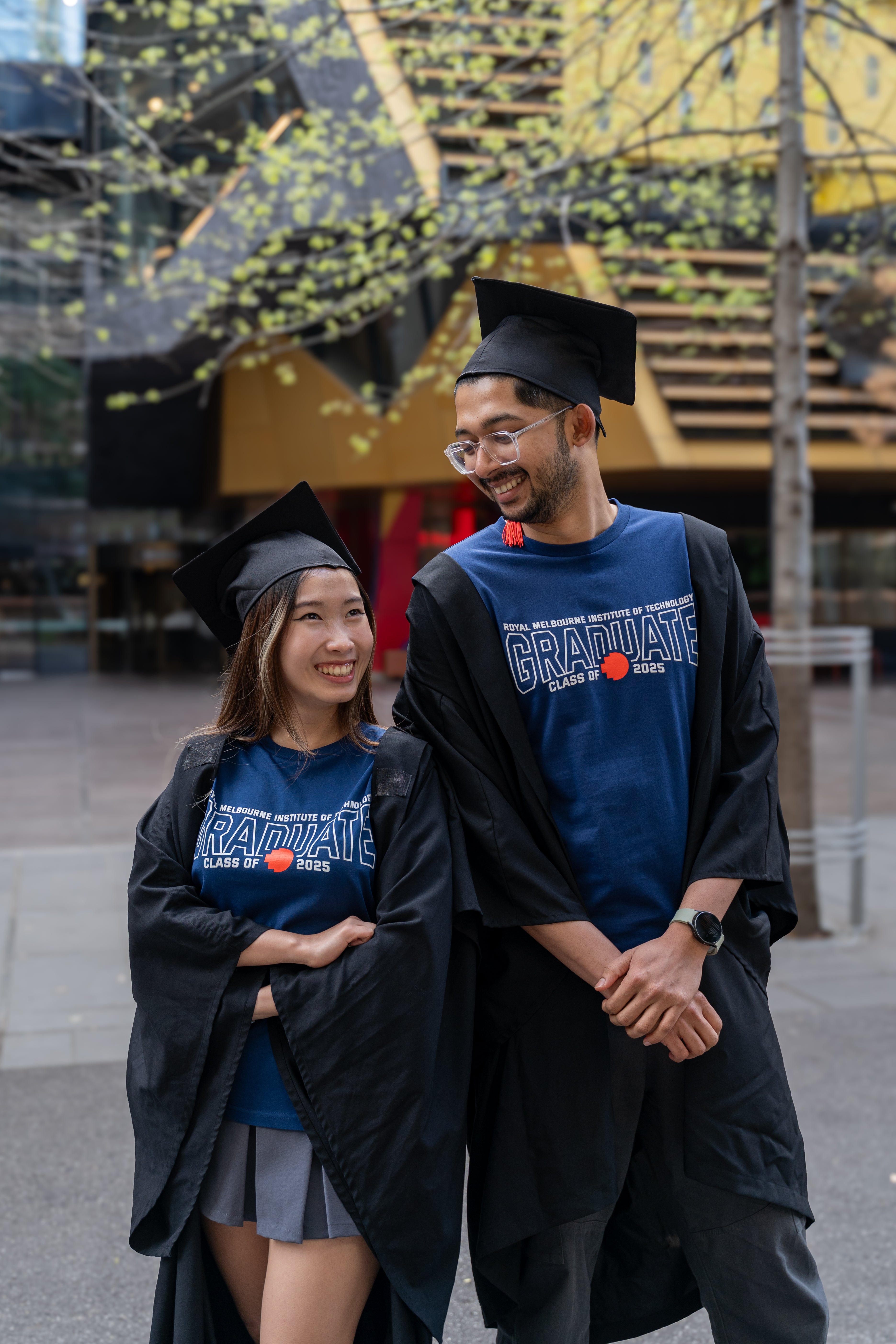 Male and female models wearing cobalt blue RMIT Class of 2025 Tees layered with black graduation gowns, both featuring bold white lettering and red pixel RMIT logos, standing outdoors on campus and smiling at each other, available at the RMIT Store.