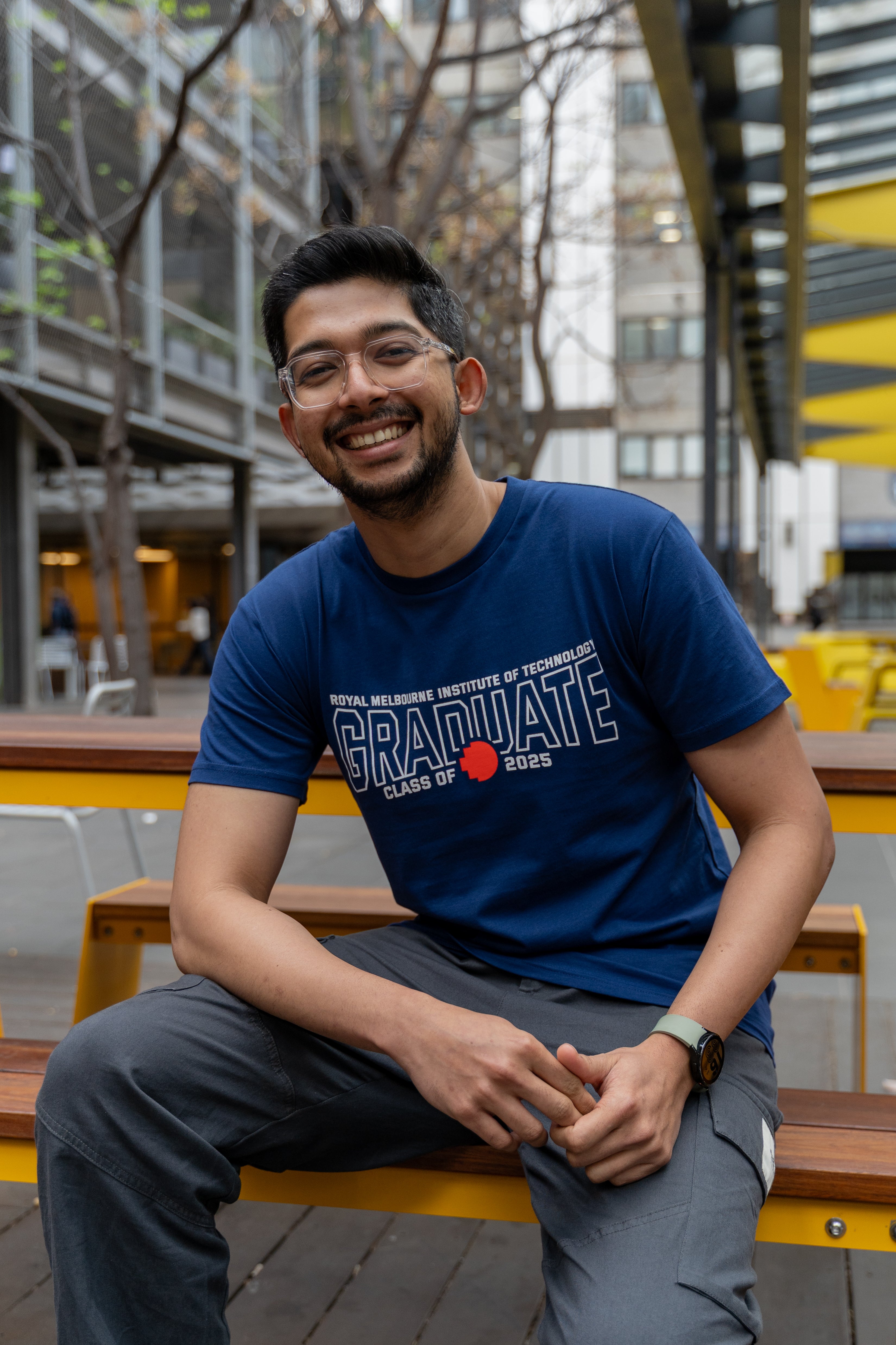 Male model wearing a cobalt blue RMIT Class of 2025 Tee with bold white “Graduate” lettering, “Royal Melbourne Institute of Technology” text above, and a red pixel RMIT logo; seated casually on an outdoor wooden bench with yellow benches on campus, smiling towards the camera, available at the RMIT Store.