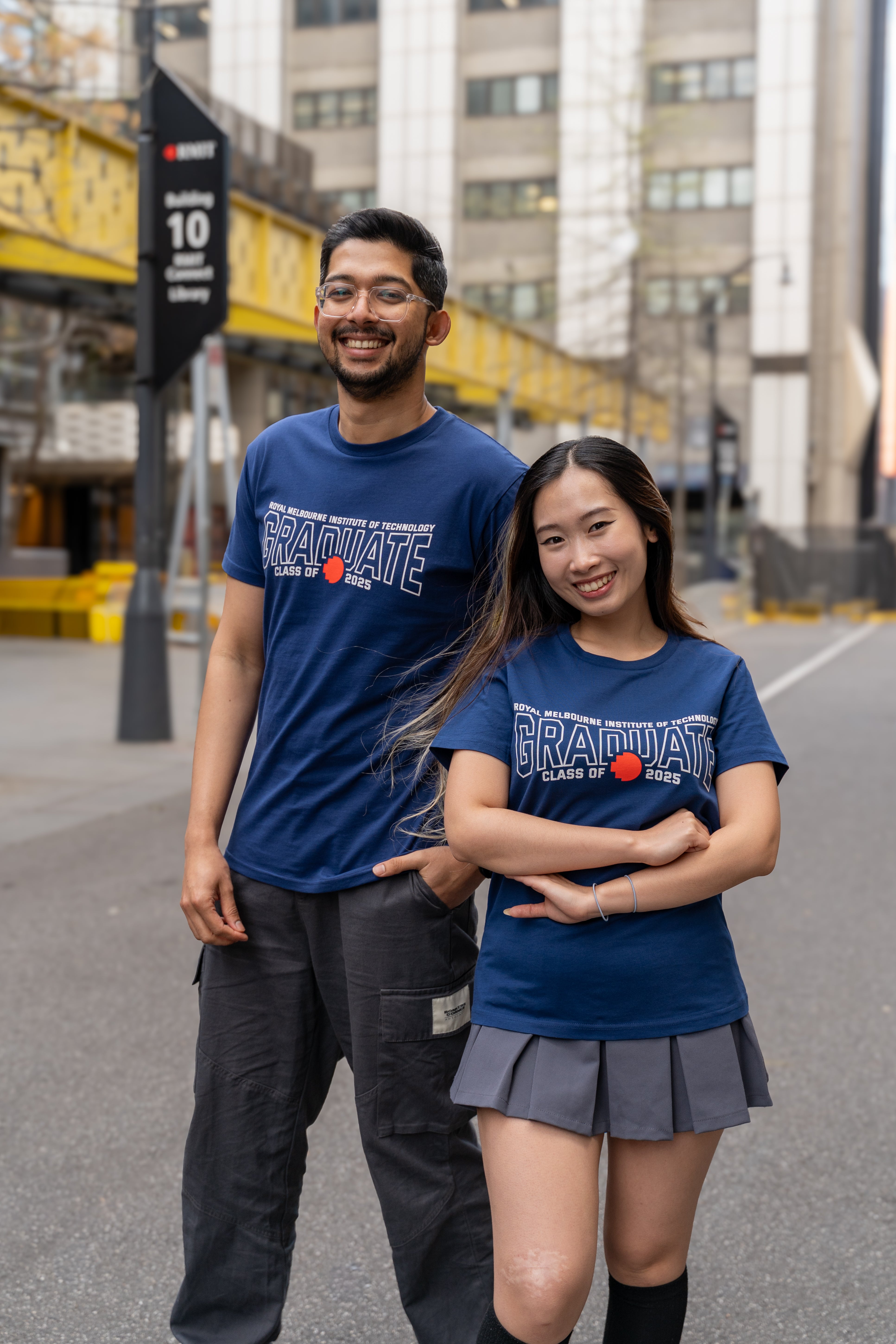 Male and female models wearing cobalt blue RMIT Class of 2025 Tees with bold white “Graduate” lettering and red pixel RMIT logos, standing confidently side by side on a city street with RMIT campus signage and yellow bridge structure in the background, available at the RMIT Store.