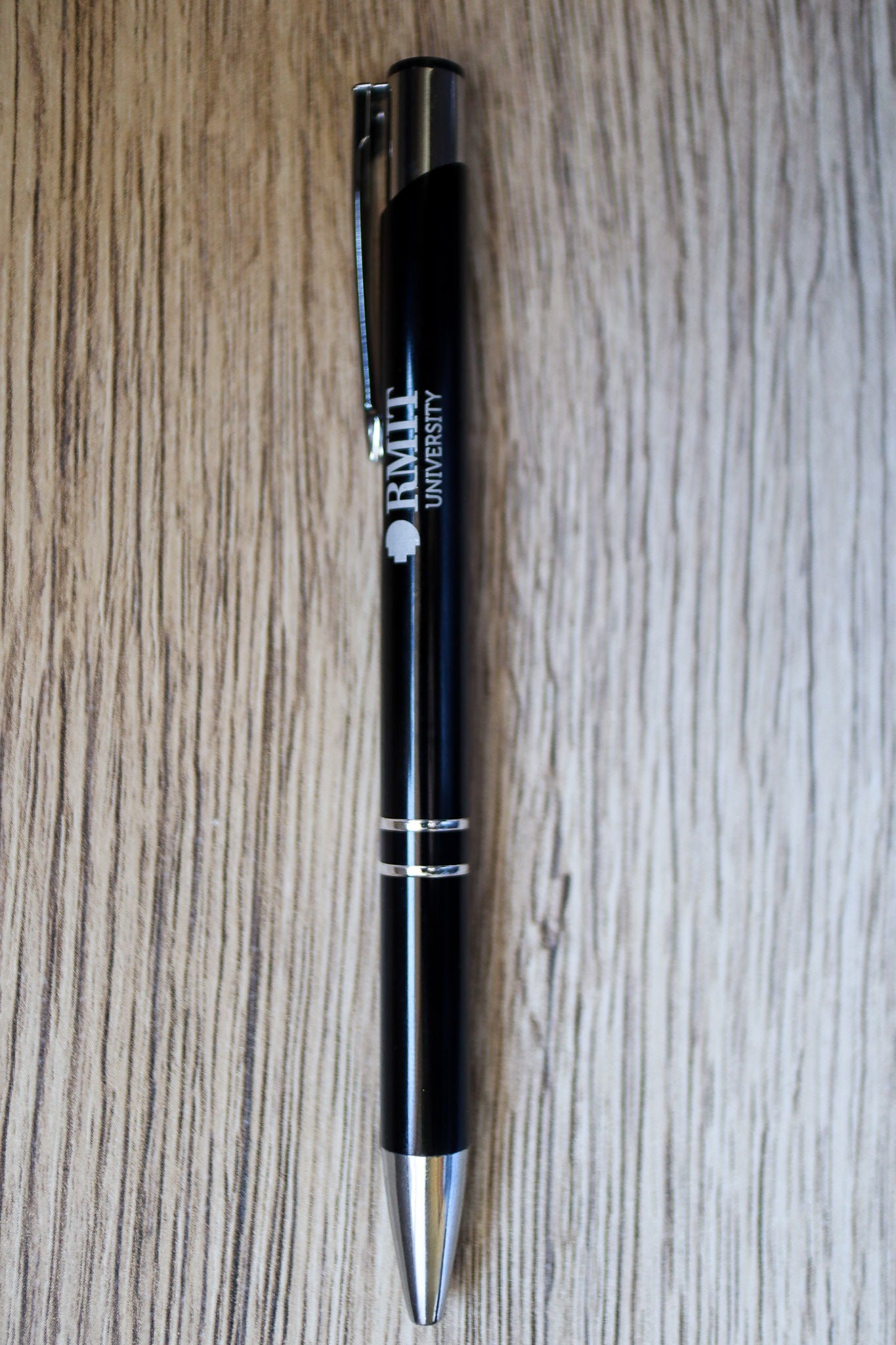 Full view of a black Classic Pen with white RMIT University branding and a silver press-end mechanism, lying flat on a natural wooden floorboard surface. Available at the RMIT Store.