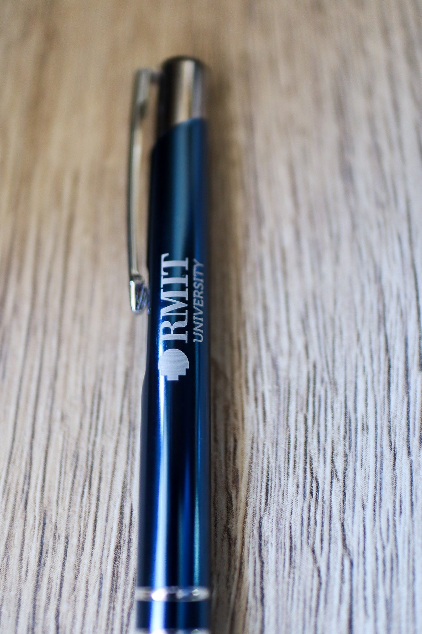 Close-up view of the bottom section of a navy Classic Pen featuring white RMIT University branding and a silver press-end mechanism. The pen rests on a natural wooden floorboard surface. Available at the RMIT Store.