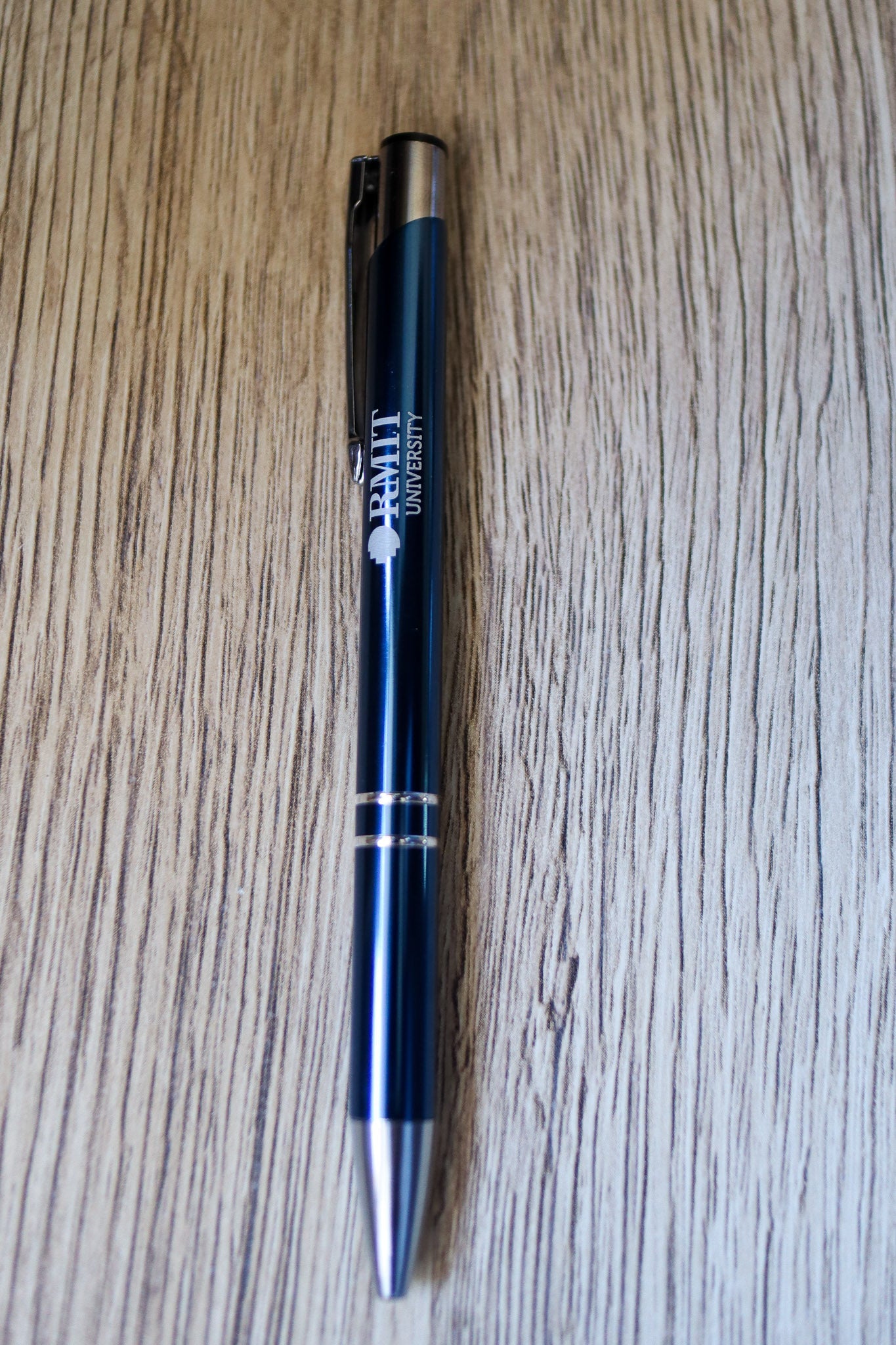 Full view of a navy Classic Pen with white RMIT University branding and a silver press-end mechanism, lying flat on a natural wooden floorboard surface. Available at the RMIT Store.