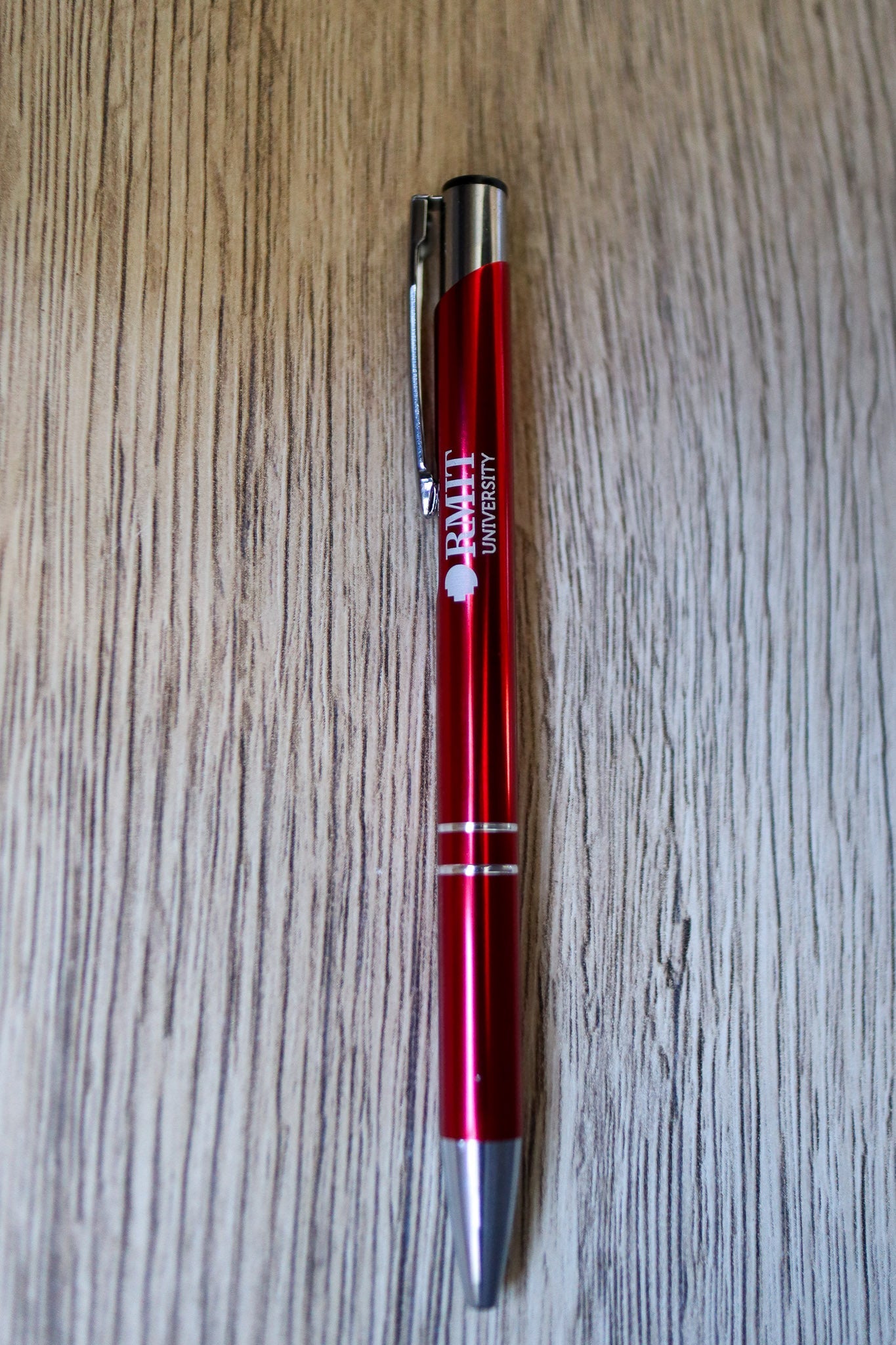 Full view of a red Classic Pen with white RMIT University branding and a silver press-end mechanism, lying flat on a natural wooden floorboard surface. Available at the RMIT Store.
