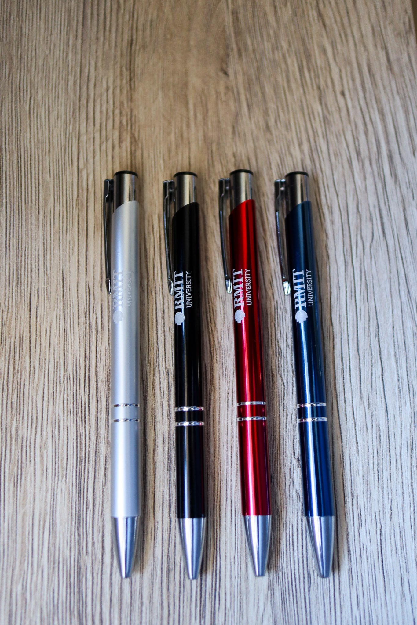 Full view of four Classic Pens in red, navy, black, and silver colours, each featuring white RMIT University branding and silver press-end mechanisms, neatly arranged side by side on a natural wooden floorboard surface. Available at the RMIT Store.
