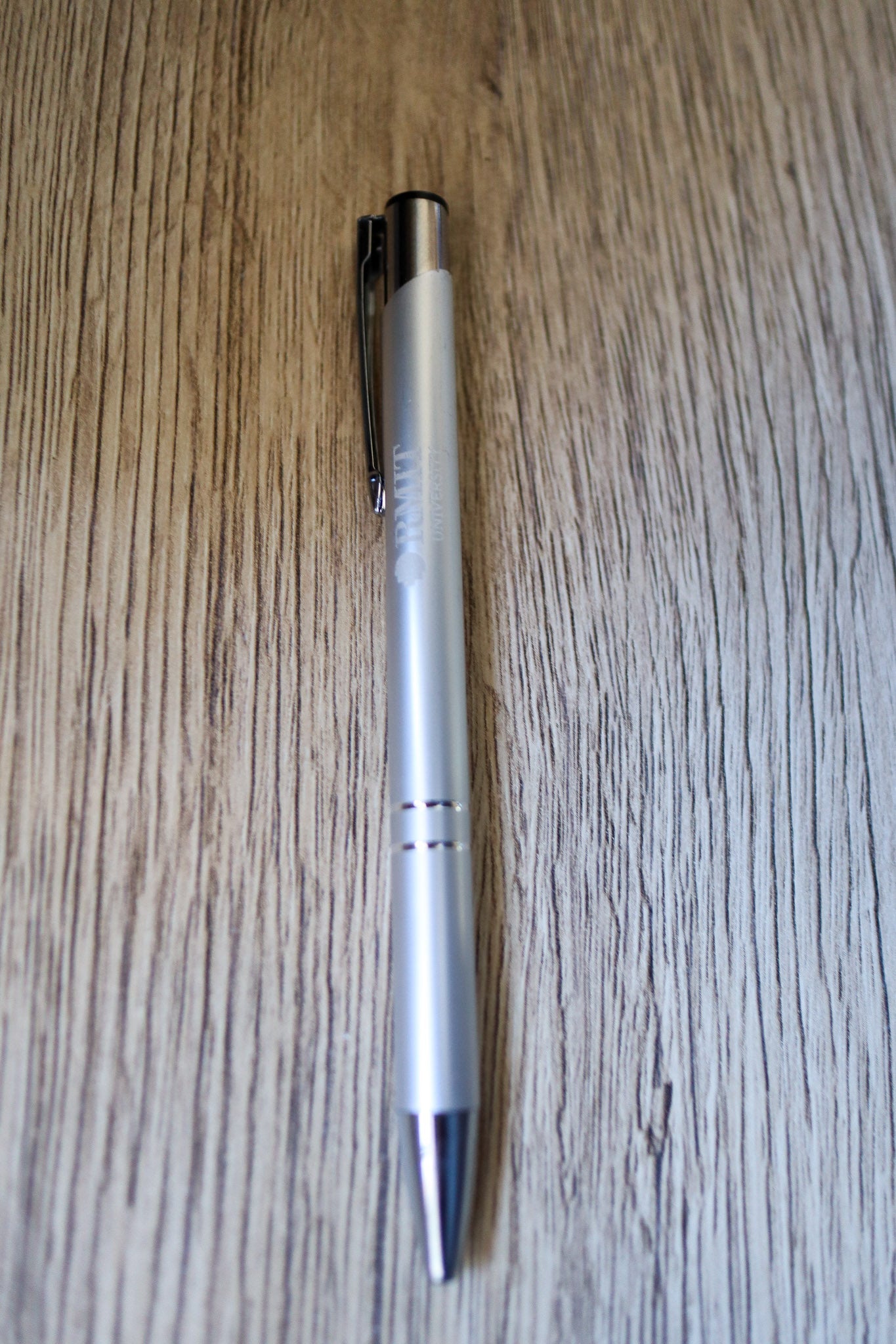 Full view of a silver Classic Pen with white RMIT University branding and a silver press-end mechanism, resting flat on a natural wooden floorboard surface. Available at the RMIT Store.