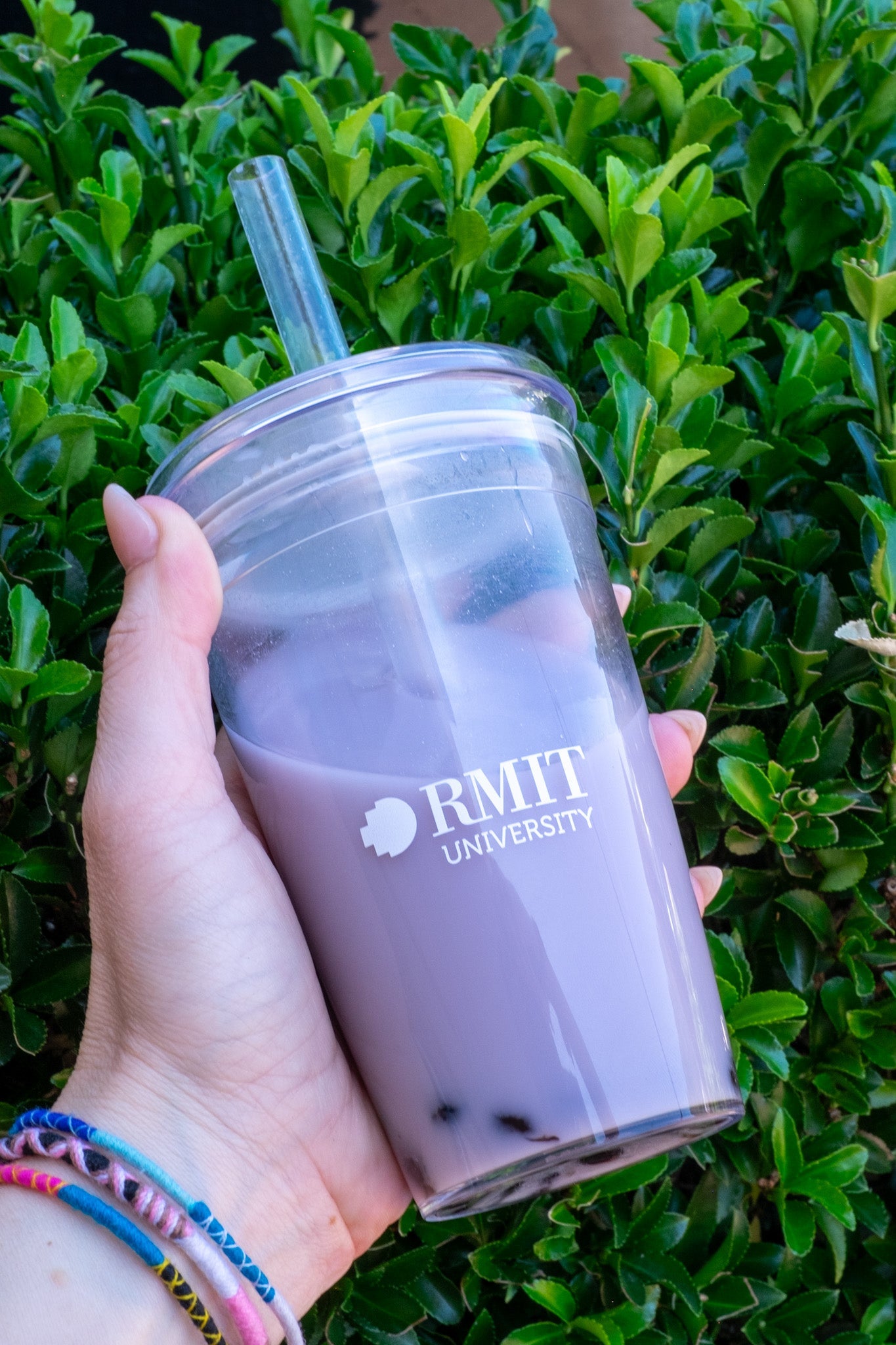Hand holding the RMIT Cold Cup with a purple beverage and tapioca pearls, against a vibrant green foliage backdrop. The clear cup displays the RMIT University logo prominently. Available through the RMIT Store.