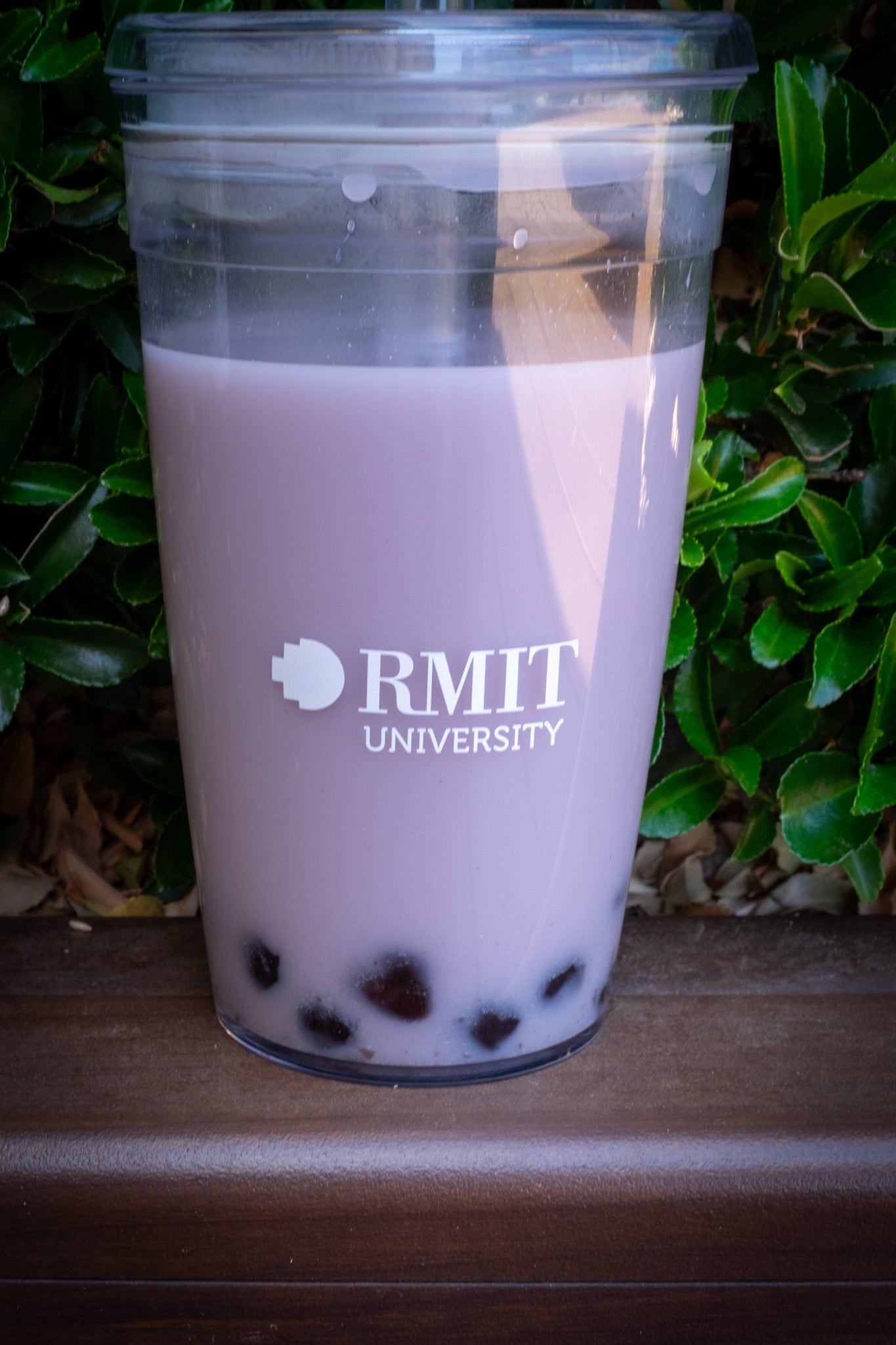 RMIT Cold Cup filled with a purple beverage and tapioca pearls, featuring the RMIT University logo. The transparent cup is placed against a backdrop of green foliage, highlighting its clear design. Available at the RMIT Store.